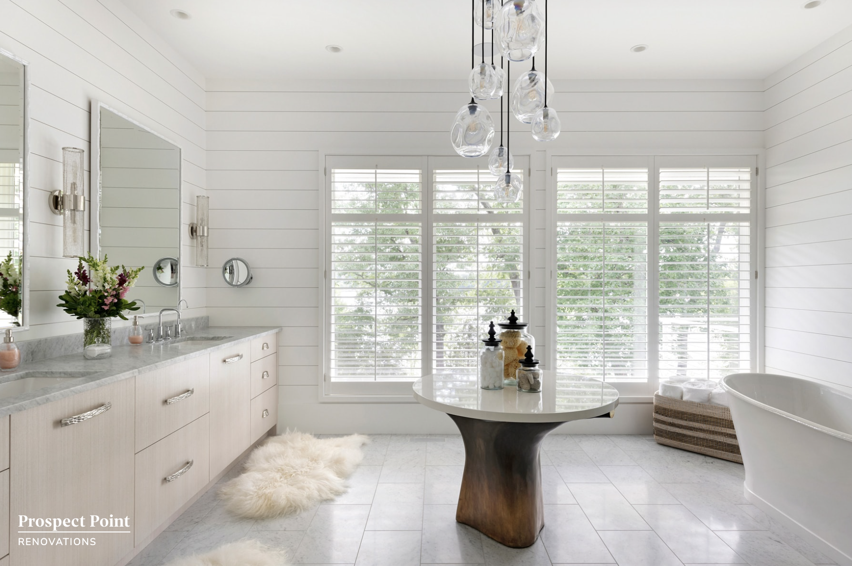 Bright bathroom with white shiplap walls and large windows with white blinds, featuring a vanity with a marble countertop, a freestanding bathtub, a round table with decorative jars, a freestanding bathtub, and a fluffy rug.