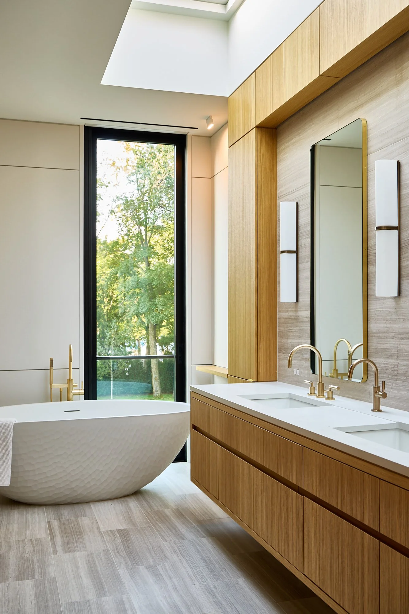 Modern bathroom with a freestanding bathtub, large window showing trees outside, and wooden cabinetry with a white countertop and gold fixtures.
