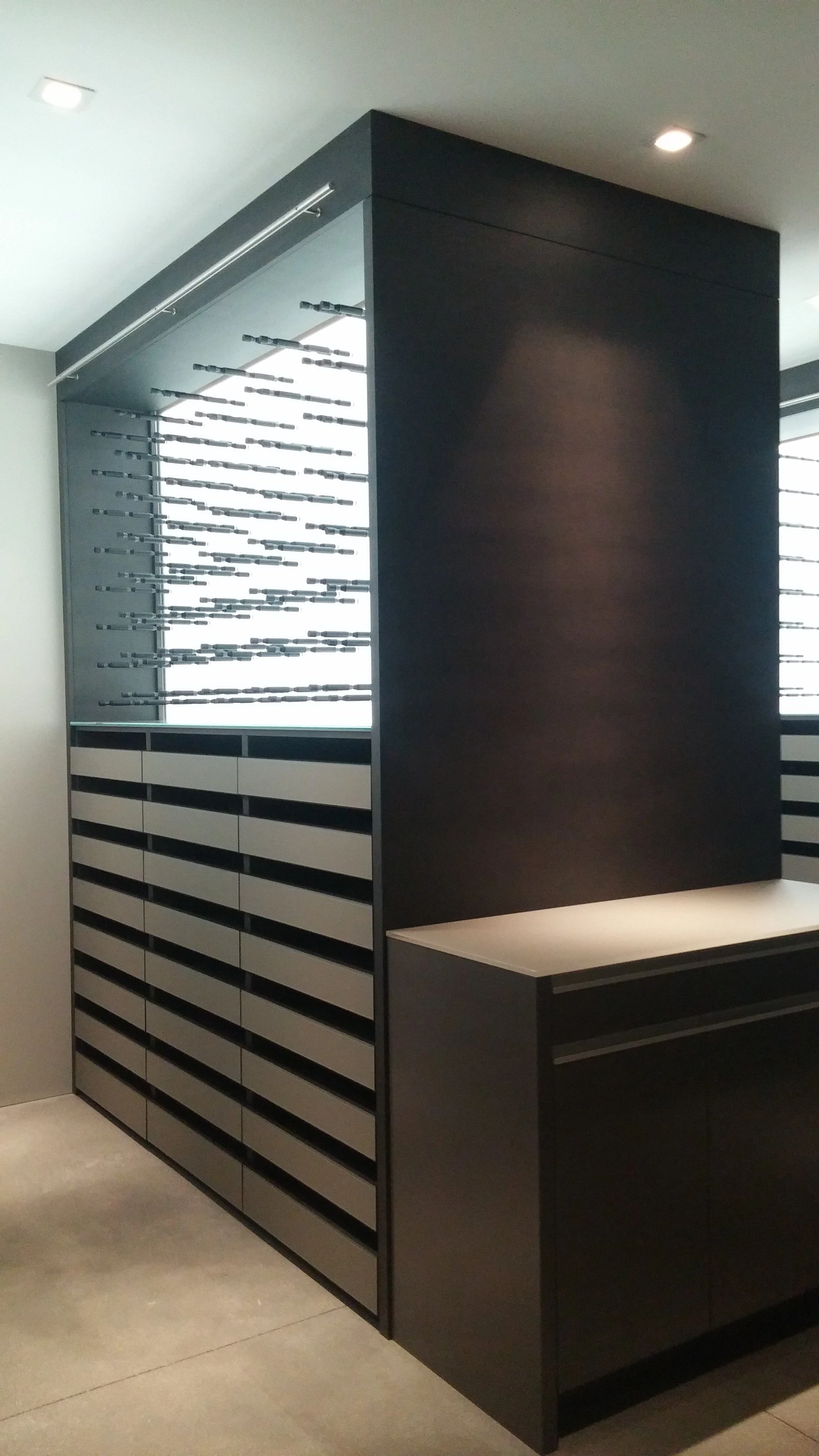A modern, black-colored wine cellar with horizontal slatted cabinets at the bottom and a built-in wine rack with horizontal holders for bottles, located in a well-lit room with white ceiling and tiled floor.