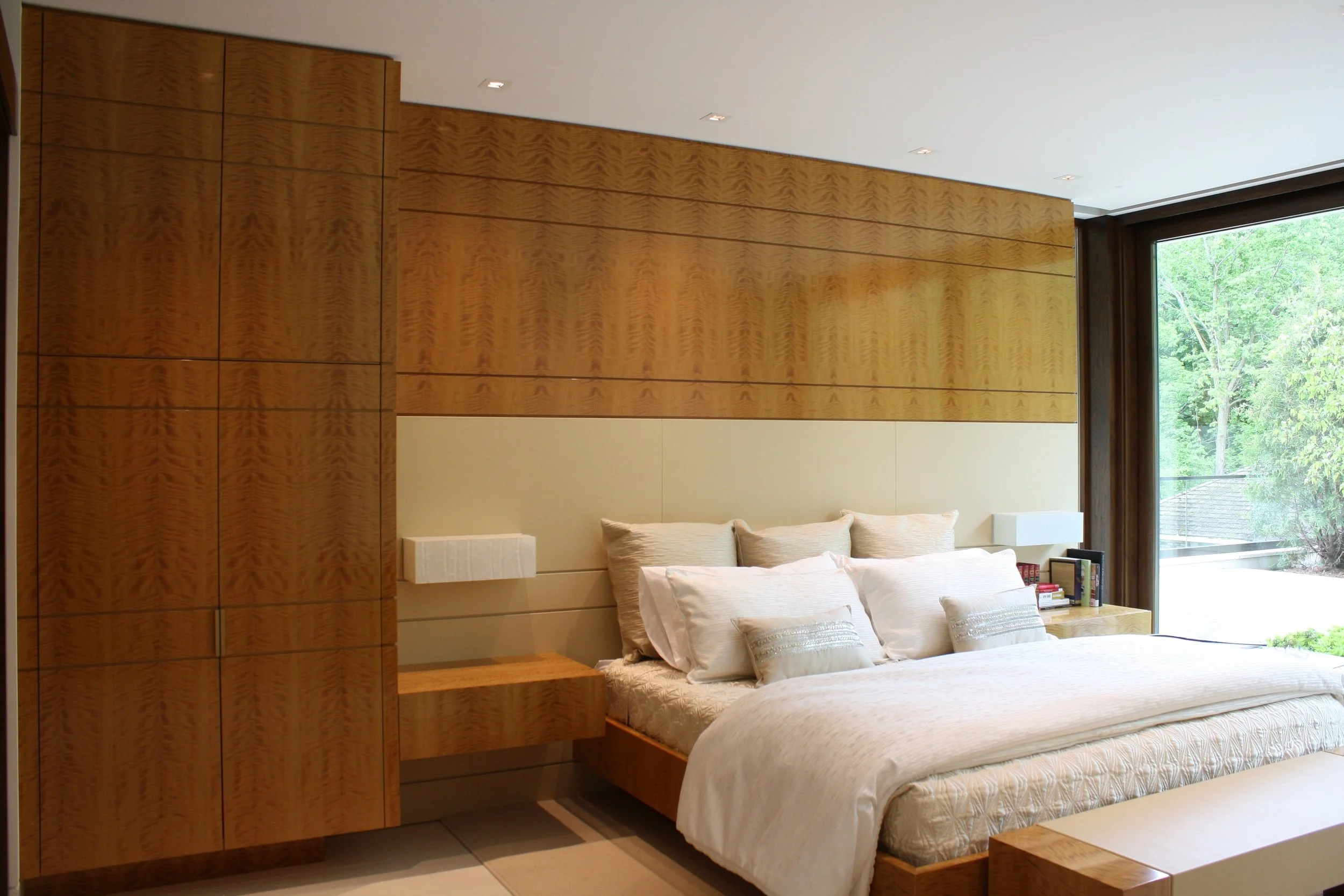 A bedroom with a large window showing green trees outside. The bed has white pillows and a beige bedspread, with a small wooden side table holding books and a remote. The wall behind the bed has a textured wood panel design, and there are small wall-