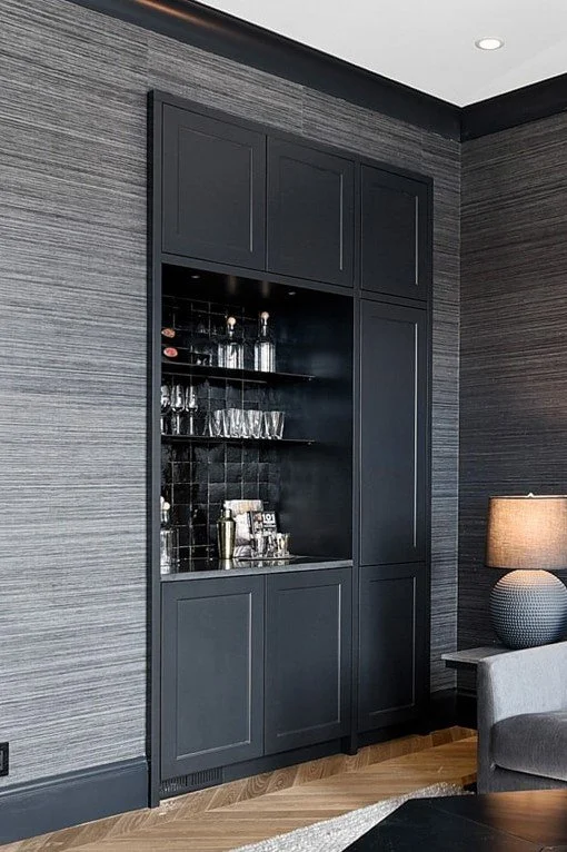 Built-in black cabinet with glass shelves holding glassware, in a modern living room with gray textured walls, a table lamp on a side table, and hardwood flooring.