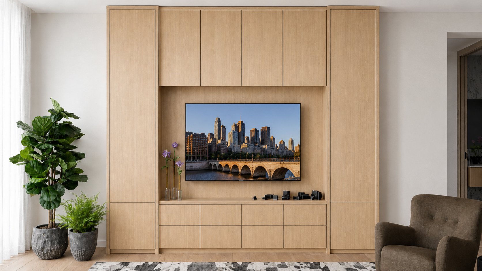Living room featuring a built-in wooden entertainment center with a flat-screen TV displaying a city skyline with a bridge over water. To the left, large potted plants with green foliage and purple flowers. To the right, a brown armchair partially vi