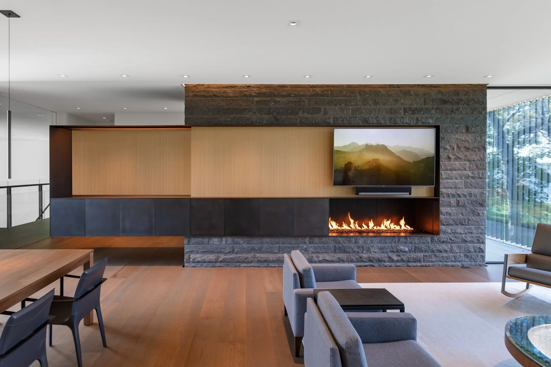 Modern living room with a stone and wood fireplace, wall-mounted TV, and large windows with curtains, decorated in neutral tones.