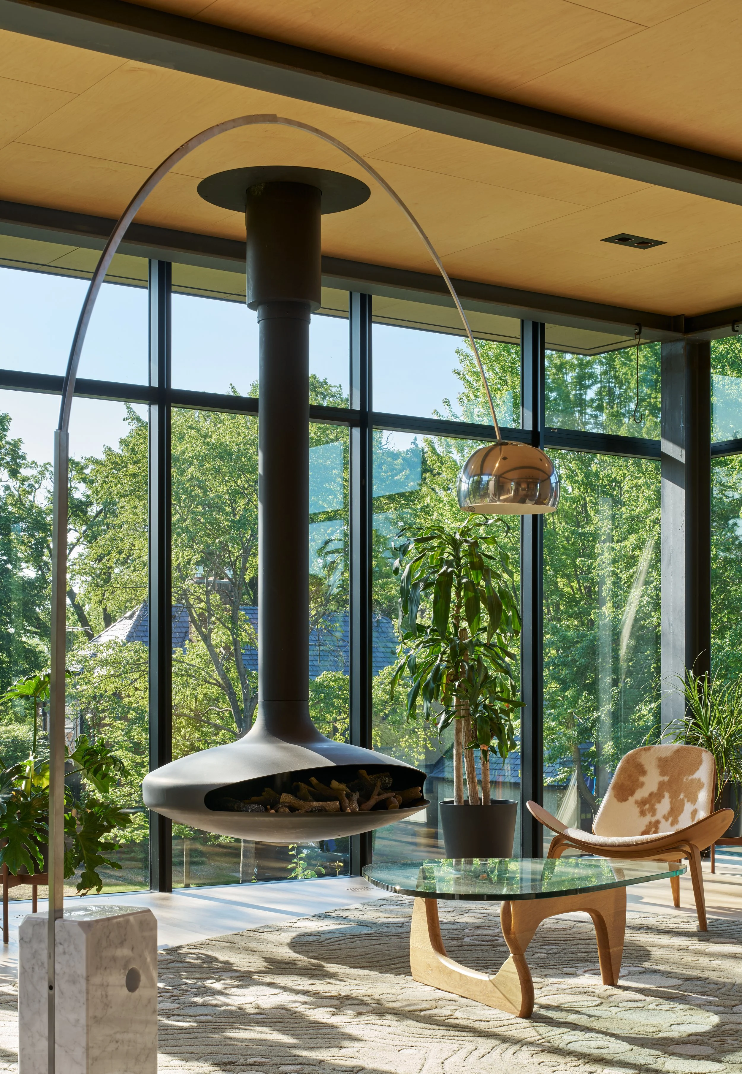Modern living room featuring large glass windows with green trees outside. A sleek, black fireplace with a curved chimney is in the center. Interior includes a glass-top wooden coffee table, a cowhide armchair, and potted plants. Natural sunlight ill