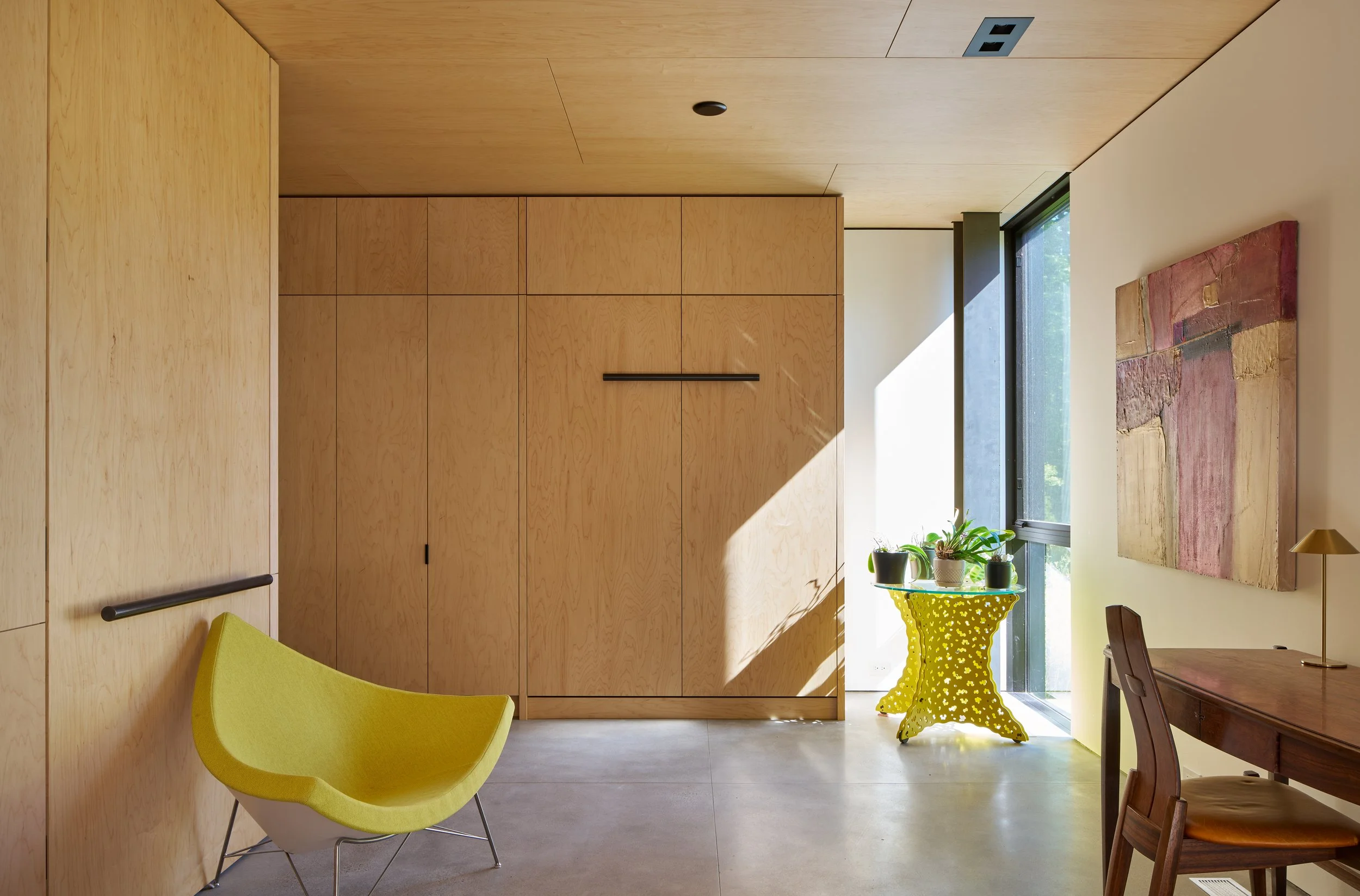 Interior room with a wooden cabinet, a yellow table with plants, a green chair, and a wooden dining table with chairs. Large window letting in natural light.