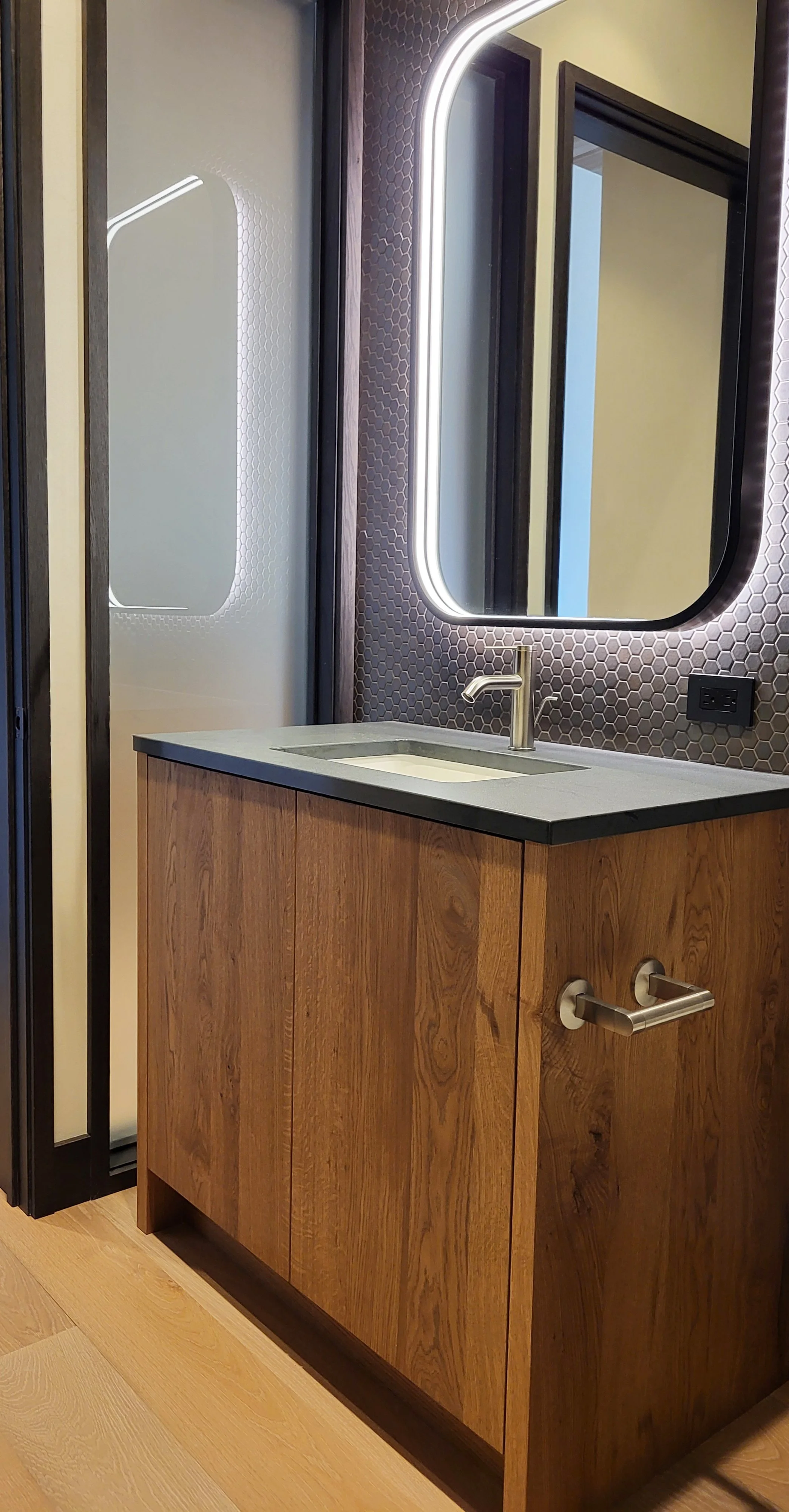A modern bathroom vanity with a wooden cabinet, black countertop, and undermount sink with a brushed metal faucet. A large mirror with LED lighting surrounds it, and there is a honeycomb patterned wall behind the mirror. A black electrical outlet is 