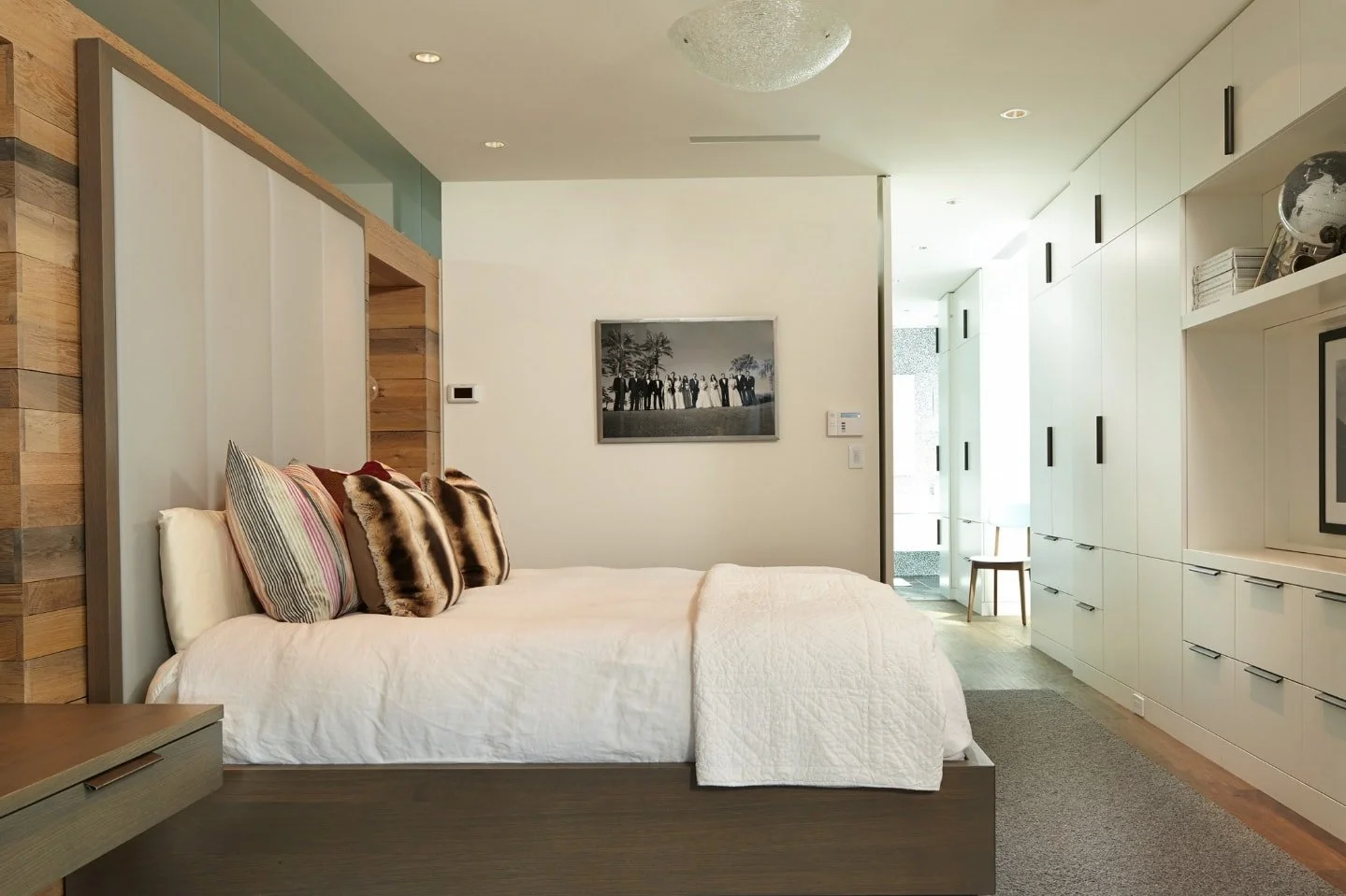Modern bedroom with a bed dressed in white linens and decorative pillows, a wall-mounted artwork, built-in white storage cabinets, and a small seating area visible through an open door.