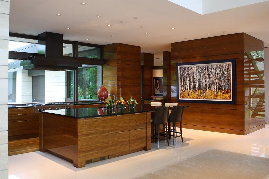 Modern kitchen with wooden cabinets, black countertops, and a large window. There is an island with a decorative vase and small plants. A big framed painting of a forest scene hangs on the wall, and there are bar stools at a small breakfast bar.