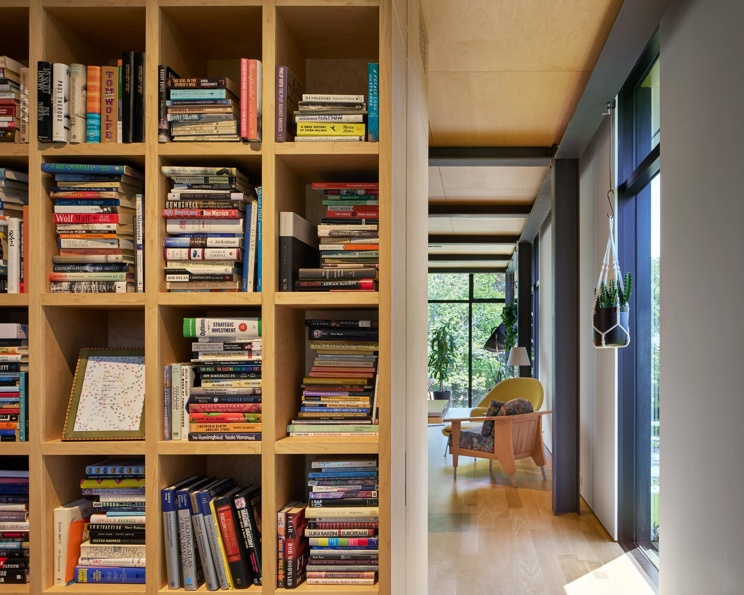 A wooden bookshelf filled with books, a framed art piece leaning against the bottom, and a cozy reading nook with a yellow armchair and plants visible through a large window.