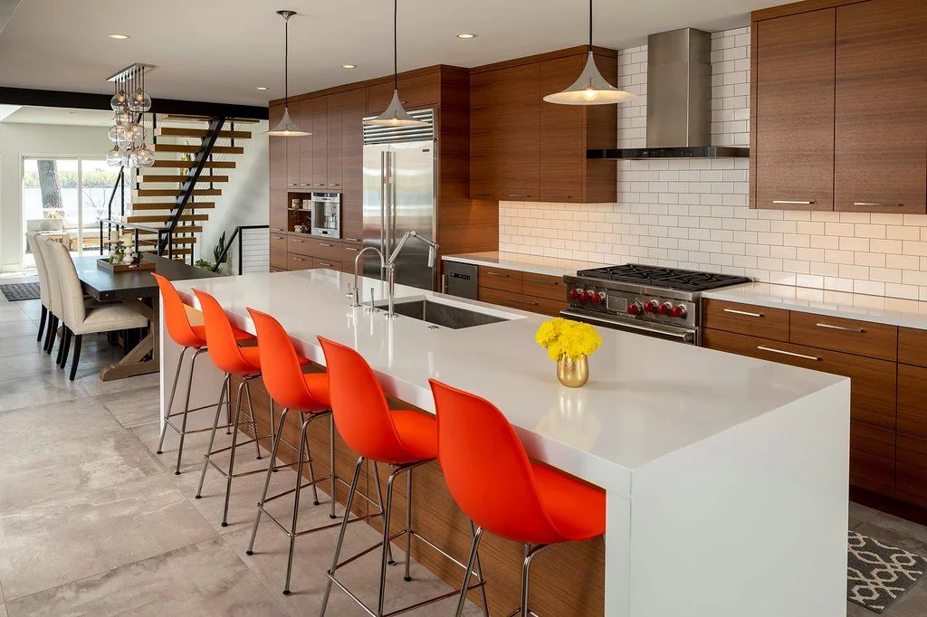 Modern kitchen with white island and red barstools, wood cabinetry, stainless steel appliances, pendant lighting, and a dining area with beige chairs and a black table in the background.