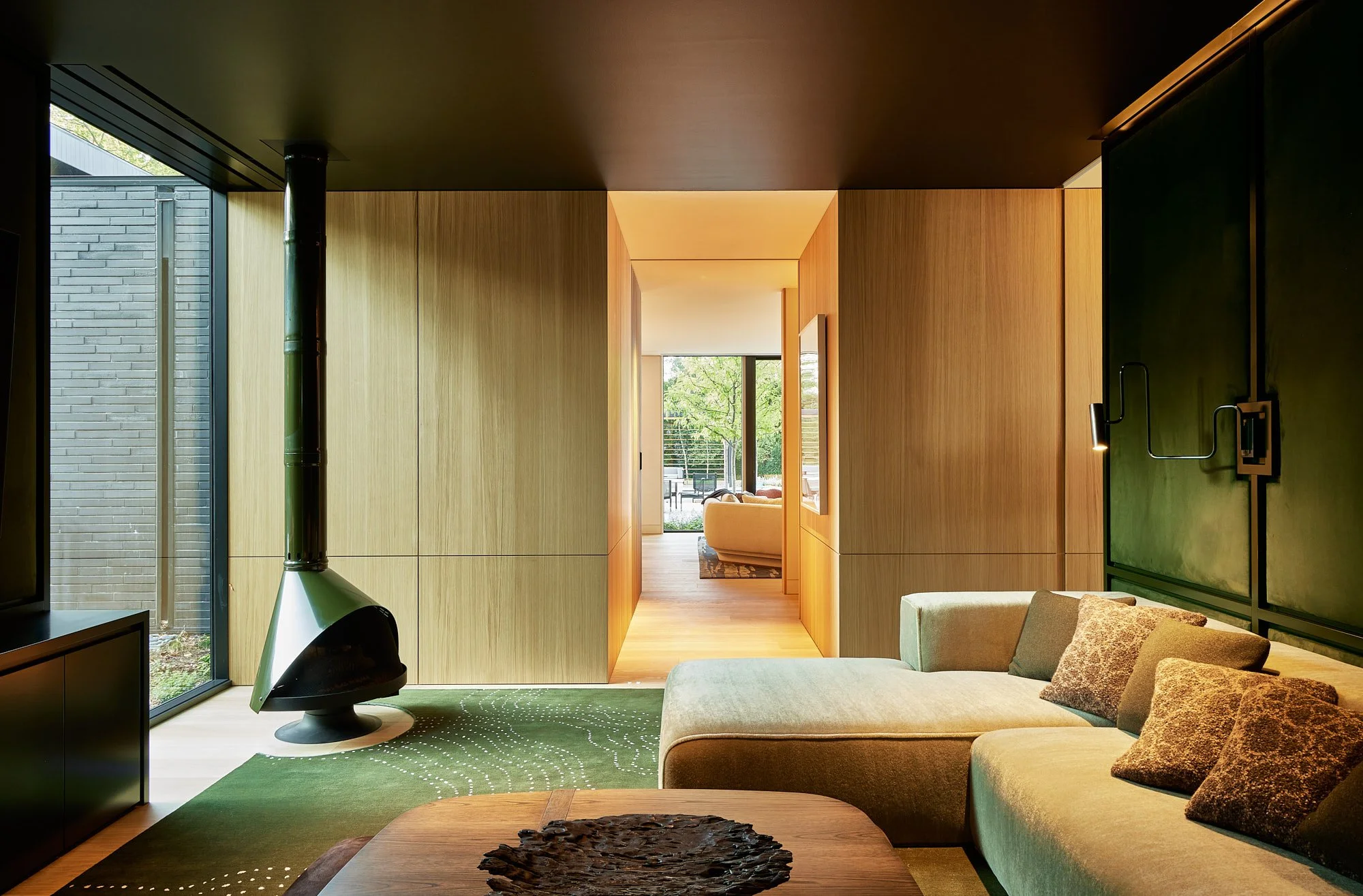 Modern living room with large green couch, wooden walls, black accent wall with a mounted TV, a fireplace, and a view through a door showing outdoor seating and greenery.