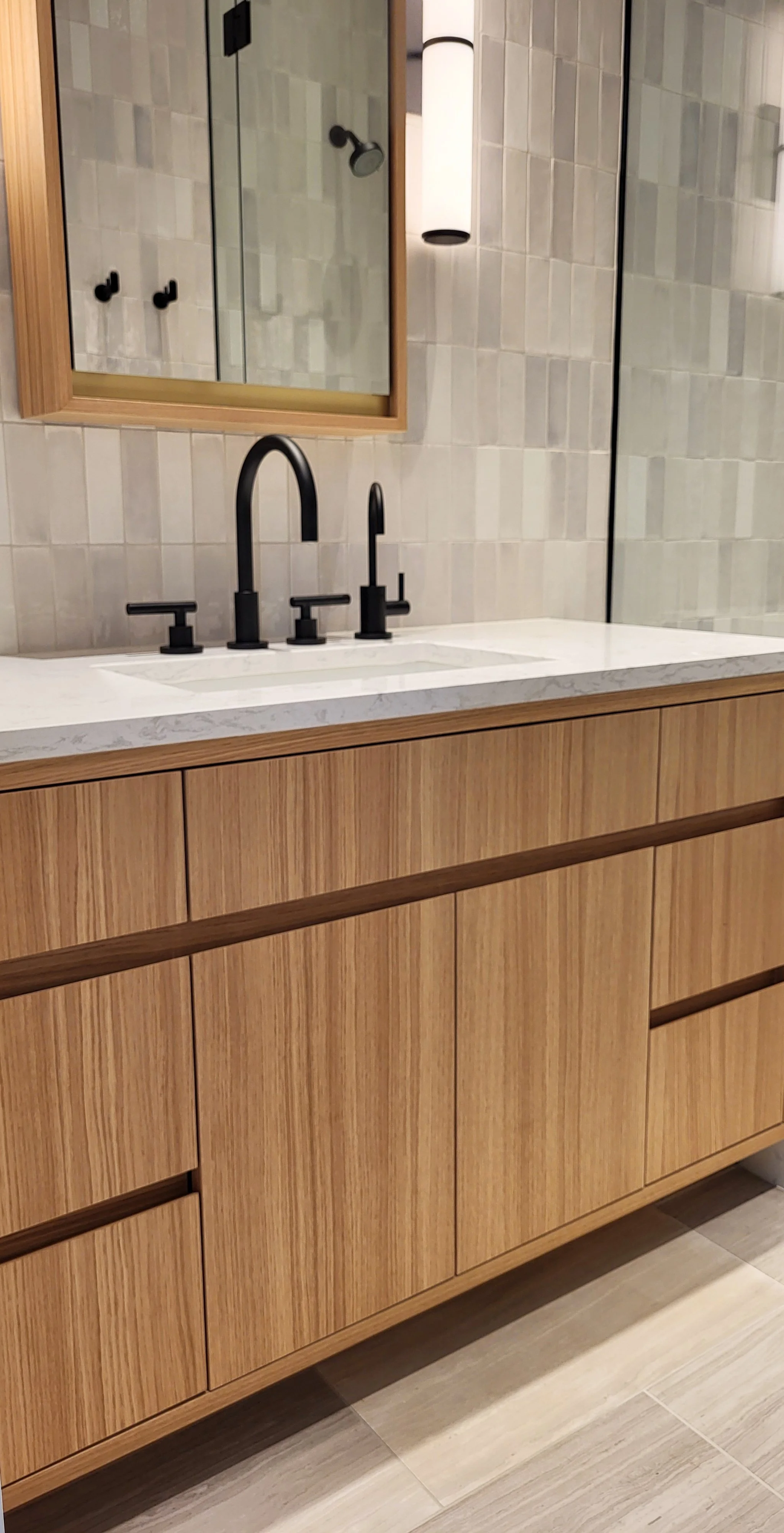 A bathroom with a wooden vanity, a white marble countertop, a black faucet with separate handles, a mirror, and a wall-mounted light fixture.