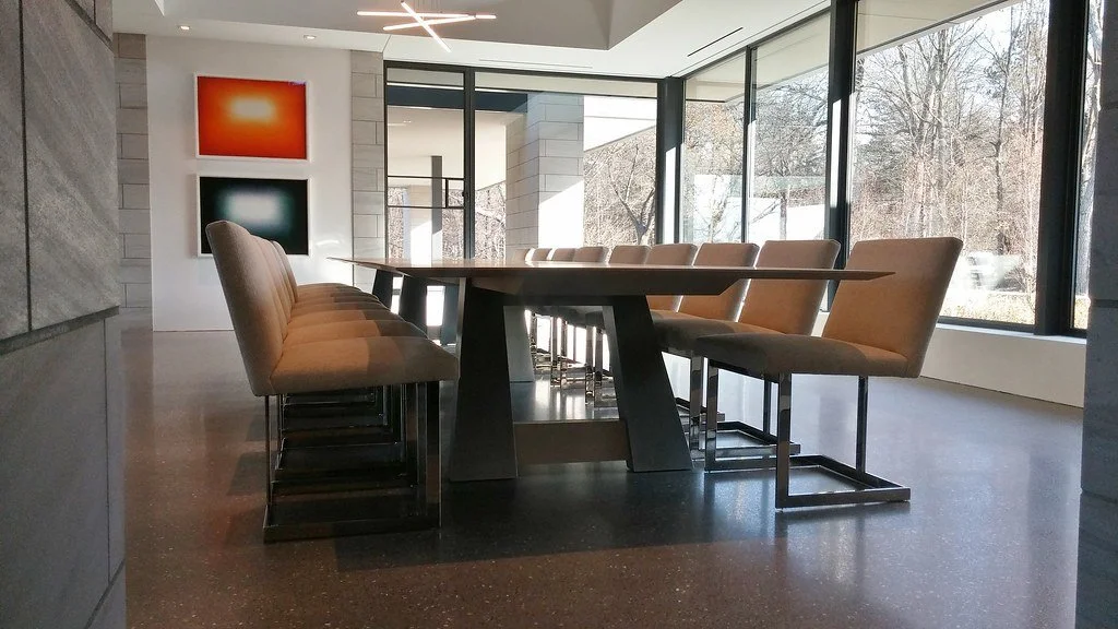 Modern indoor dining area with a long rectangular table and beige chairs, large windows overlooking a wooded area, and abstract artwork on the wall.