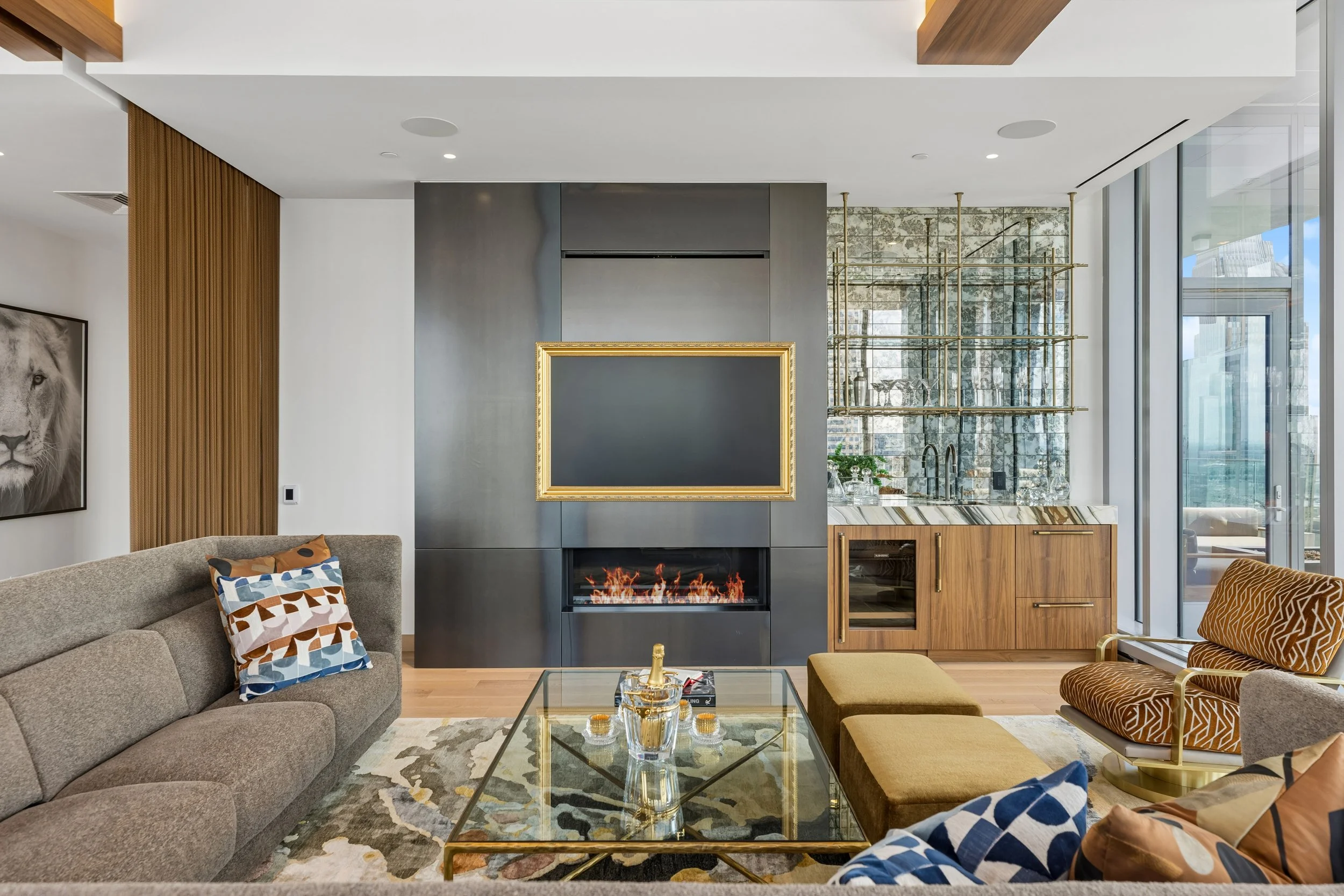 Modern living room with a gray couch, mustard and patterned pillows, glass coffee table, and a fireplace with a dark gray surround. There is a large picture of a lion on the white wall, wooden accents, and glass doors leading outside. A gold-framed m