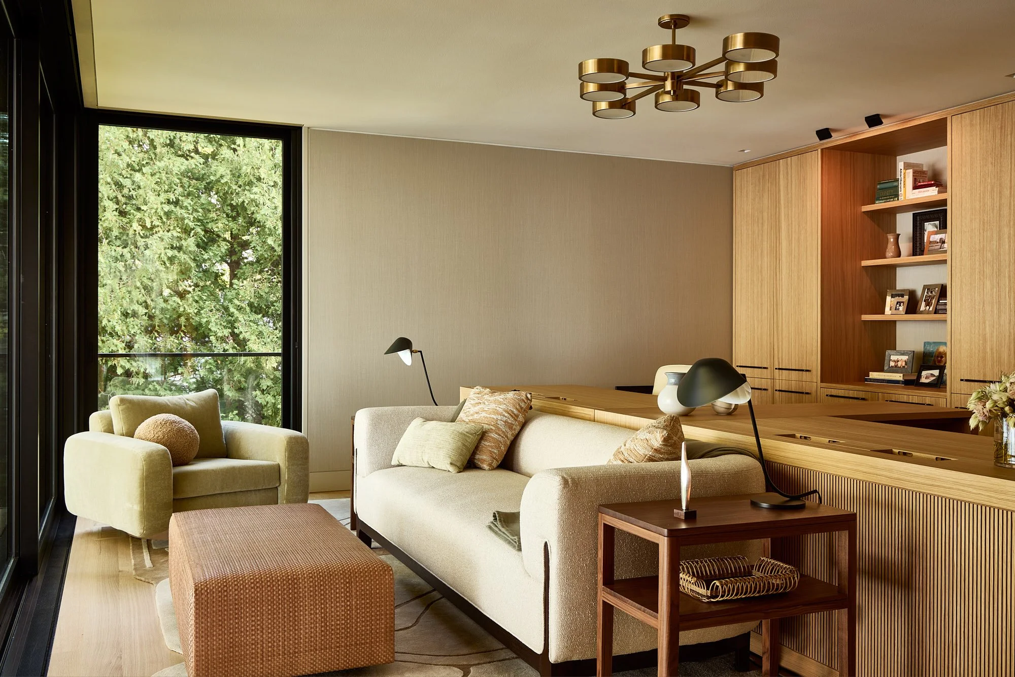 Living room with cream sofa, armchair, end table, and modern wooden shelving unit with books and framed photos, large window with green trees outside, and contemporary chandelier on ceiling.