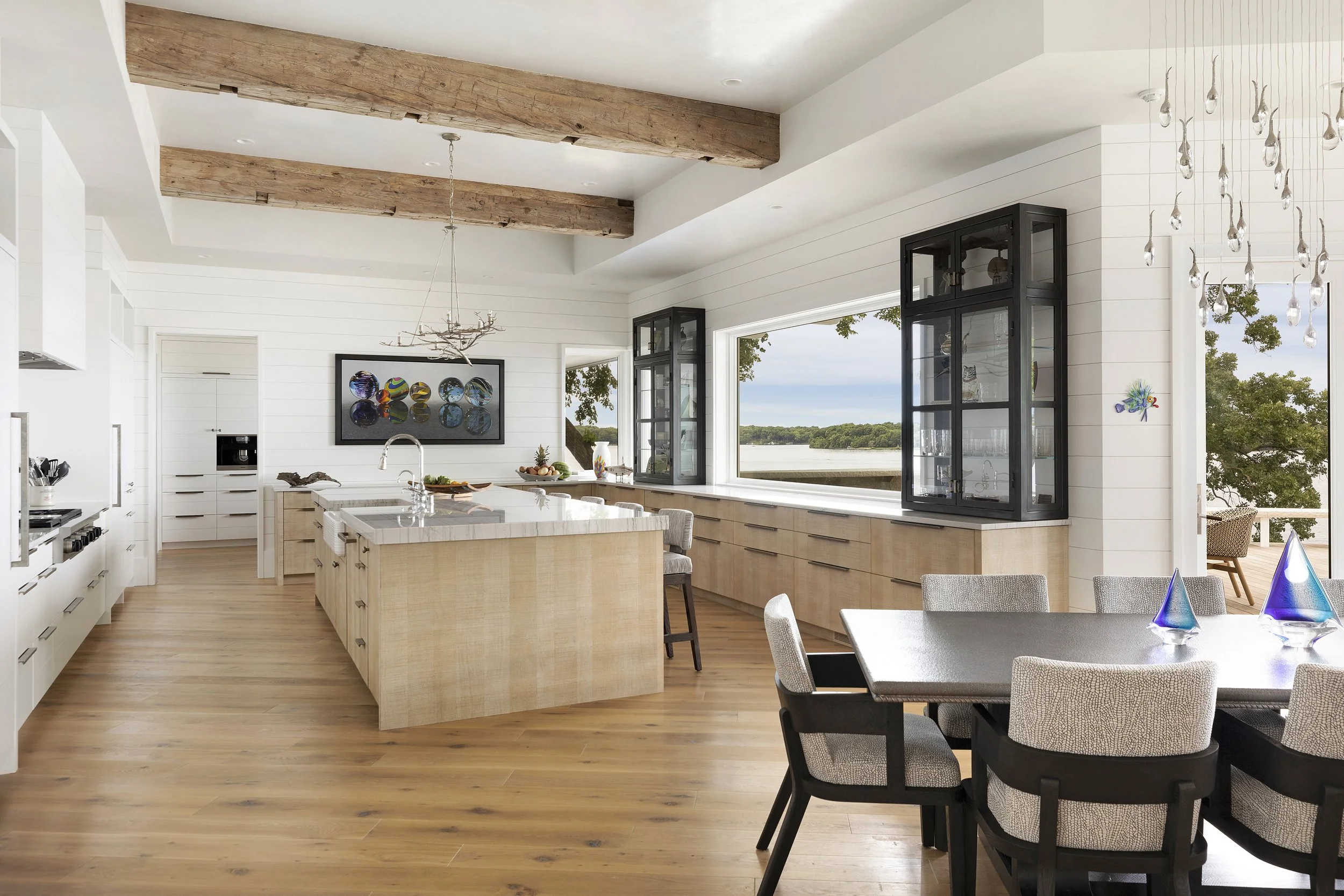 Modern kitchen with white cabinetry, light wood island, and large windows overlooking a body of water with trees. A dining table with upholstered chairs is in the foreground, and contemporary glass and wood decor accents are visible.