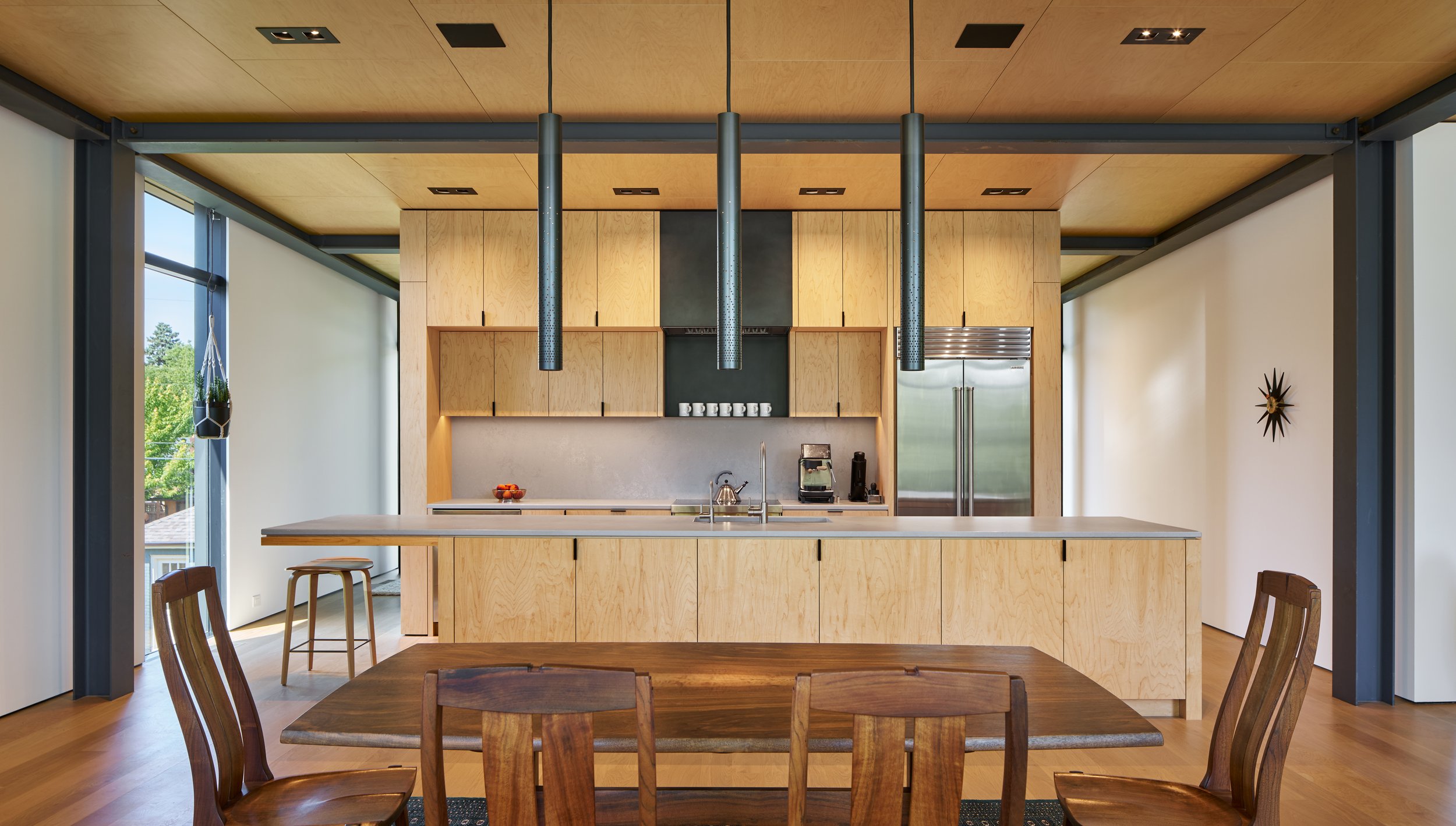 Modern kitchen with wooden cabinets, a kitchen island, and black pendant lights hanging from a wood ceiling. There are large windows, chairs, a stainless steel refrigerator, and minimal decor.