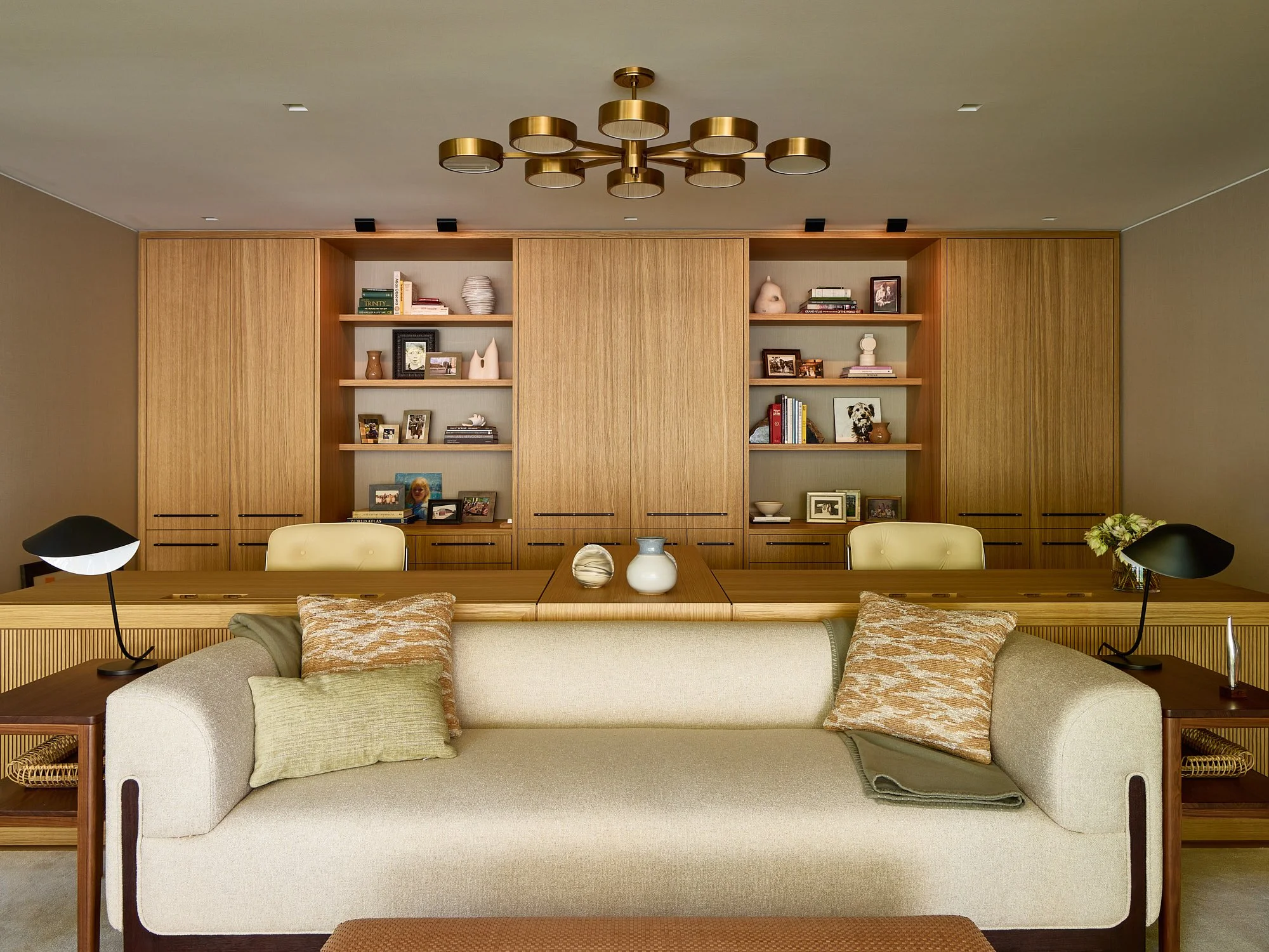 Living room with a beige sofa, green and beige pillows, wooden coffee table with vases, black and white lamps on side tables, and a wooden wall with open shelves displaying books, framed photos, and decorative objects.
