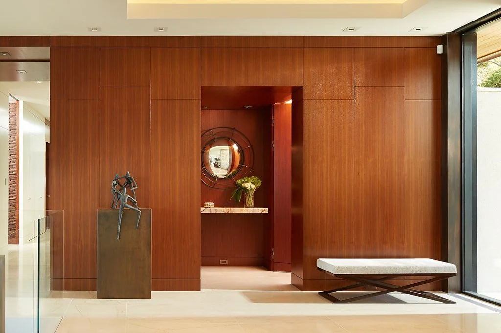Modern interior with a wooden accent wall, a sculpture on a pedestal, a decorative round mirror, a marble console with a vase of flowers, and a bench near a large window.