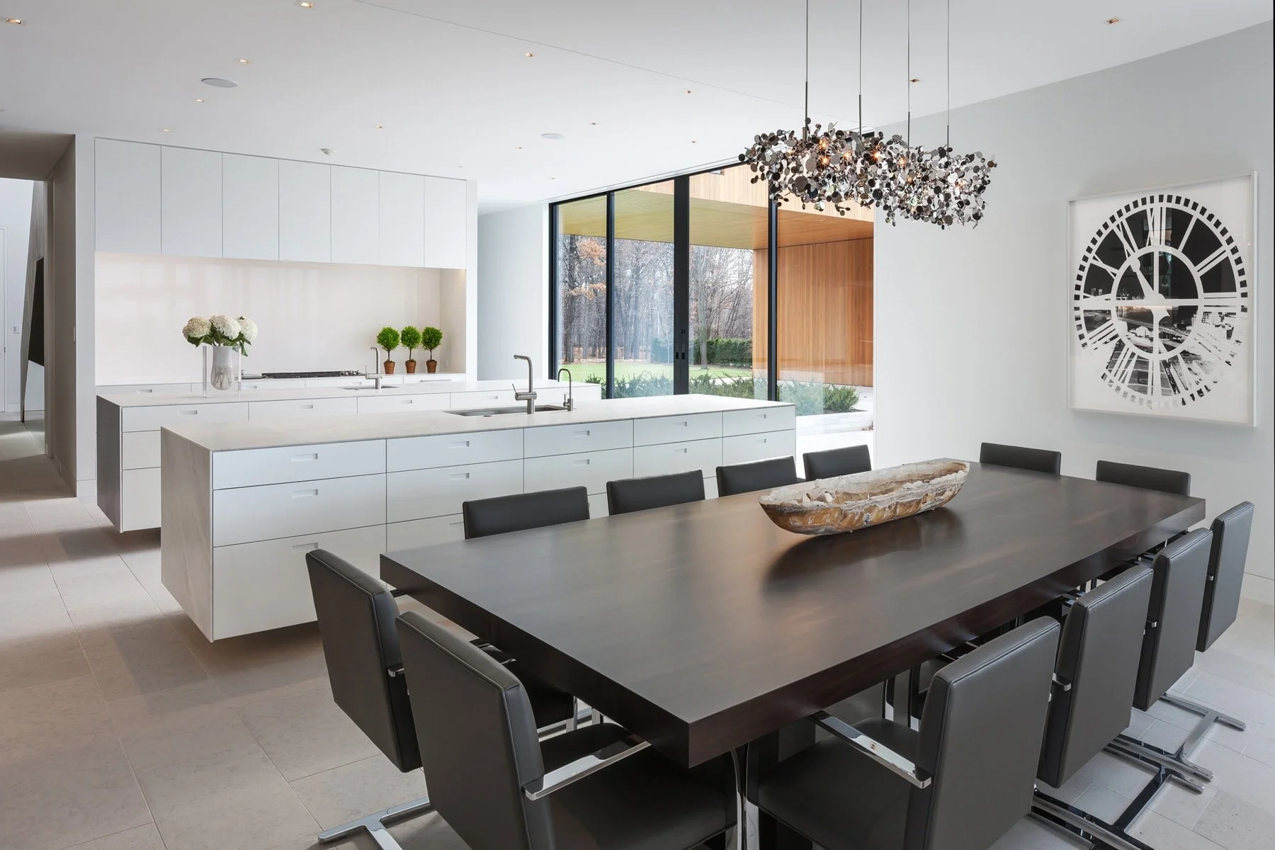 Modern dining room with a large dark wood table surrounded by black chairs, a decorative bowl on the table, a white kitchen island with a sink, and large floor-to-ceiling windows offering a view of the outdoors.