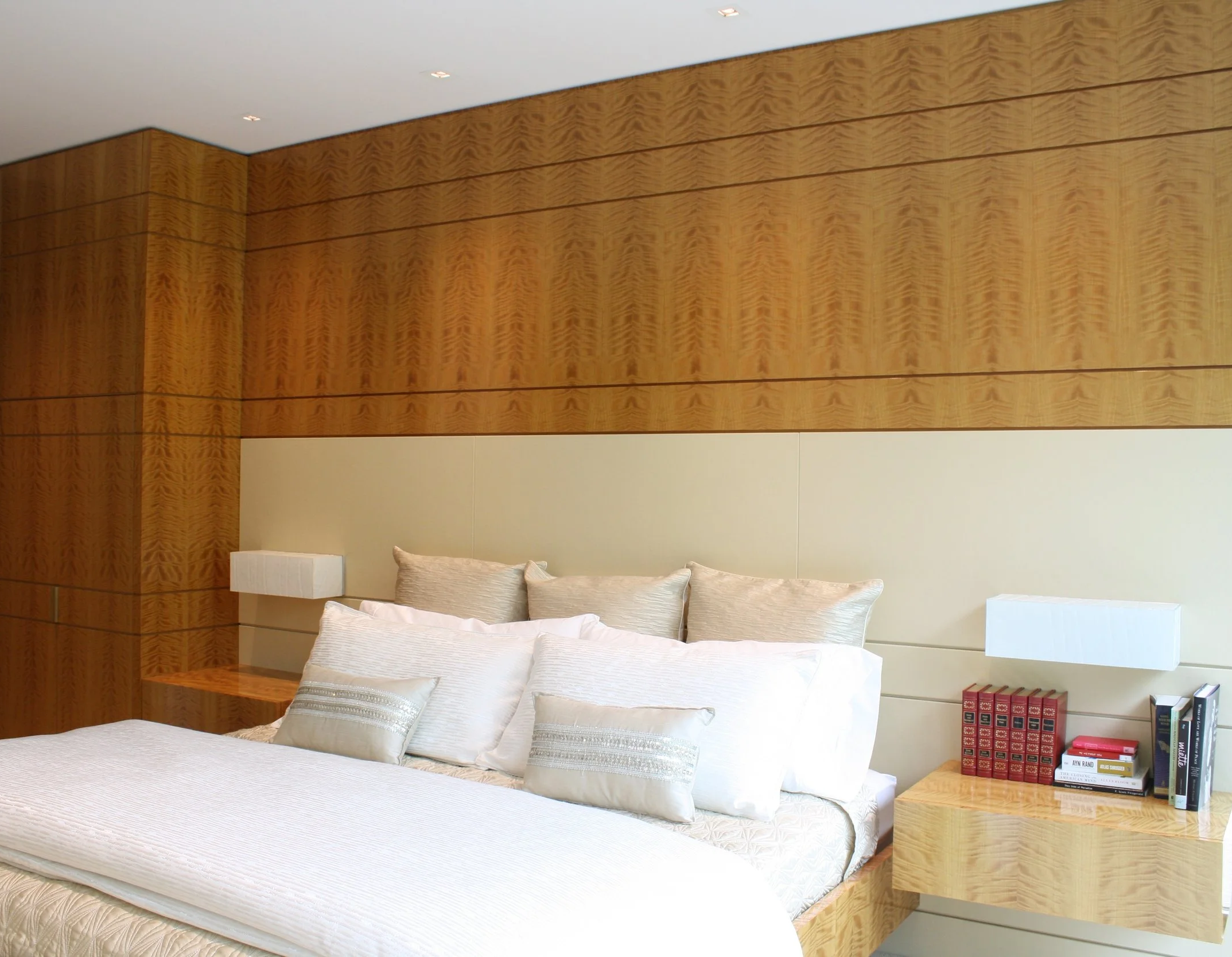 A modern bedroom with a white bed, decorative pillows, a wooden bedside table with books, a beige wall, and a textured wooden headboard extending to the ceiling.