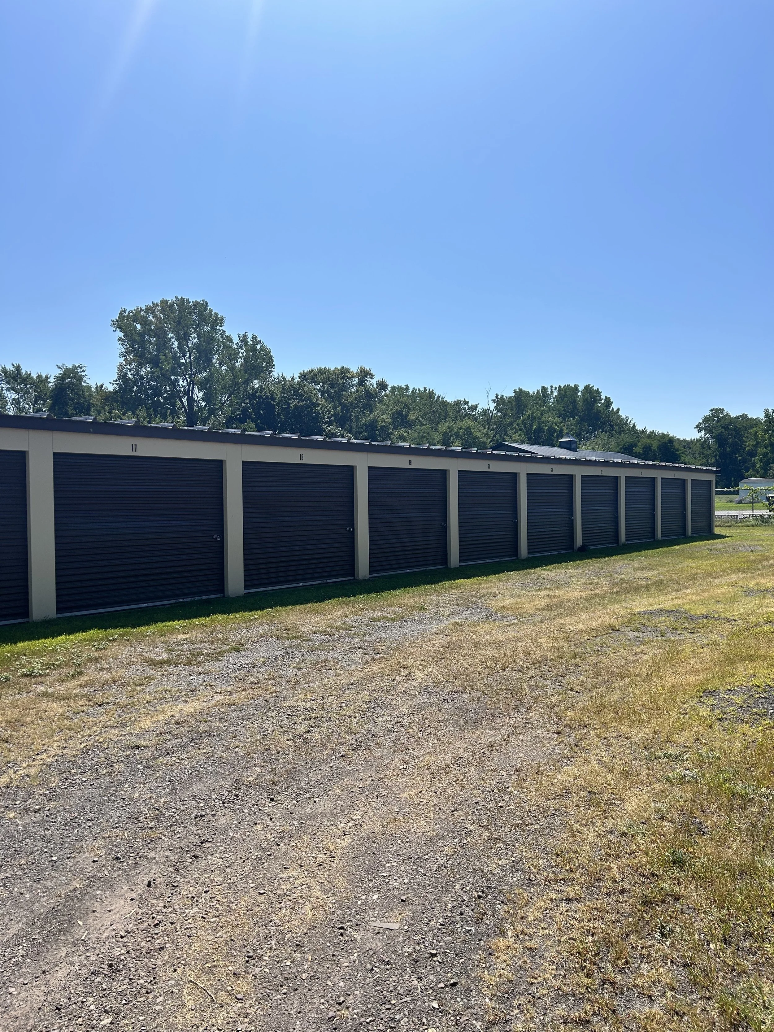 Row of storage units with black doors and white frames, numbered 17 to 20, on a gravel lot with grass patches, under a clear blue sky.