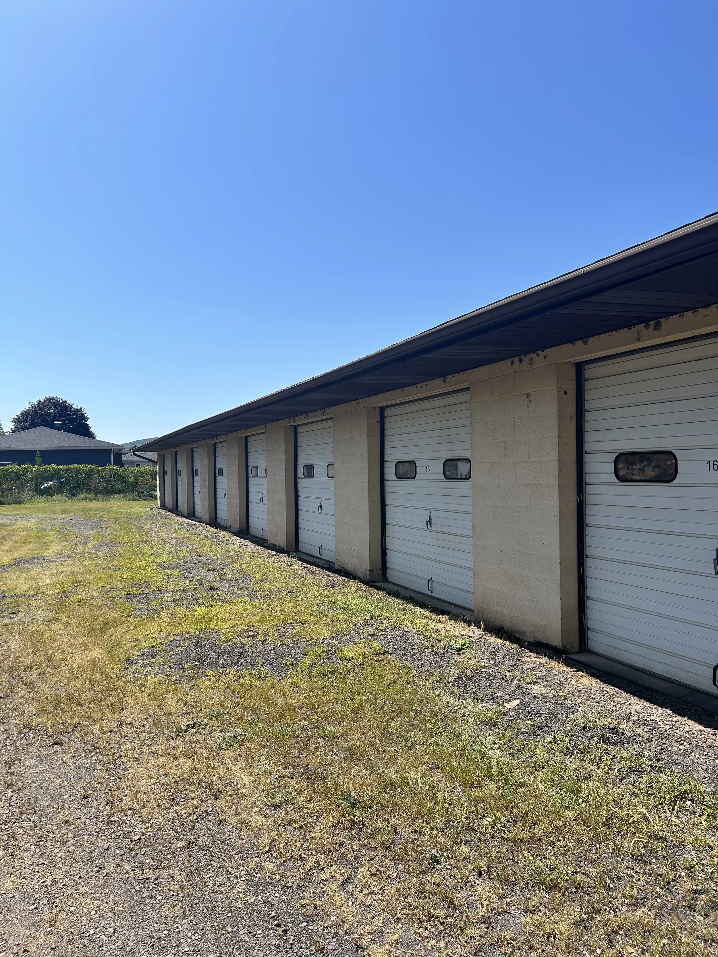Row of storage unit garages with white roll-up doors and small windows, under a sloped roof, on a grassy and gravel lot with a clear blue sky.