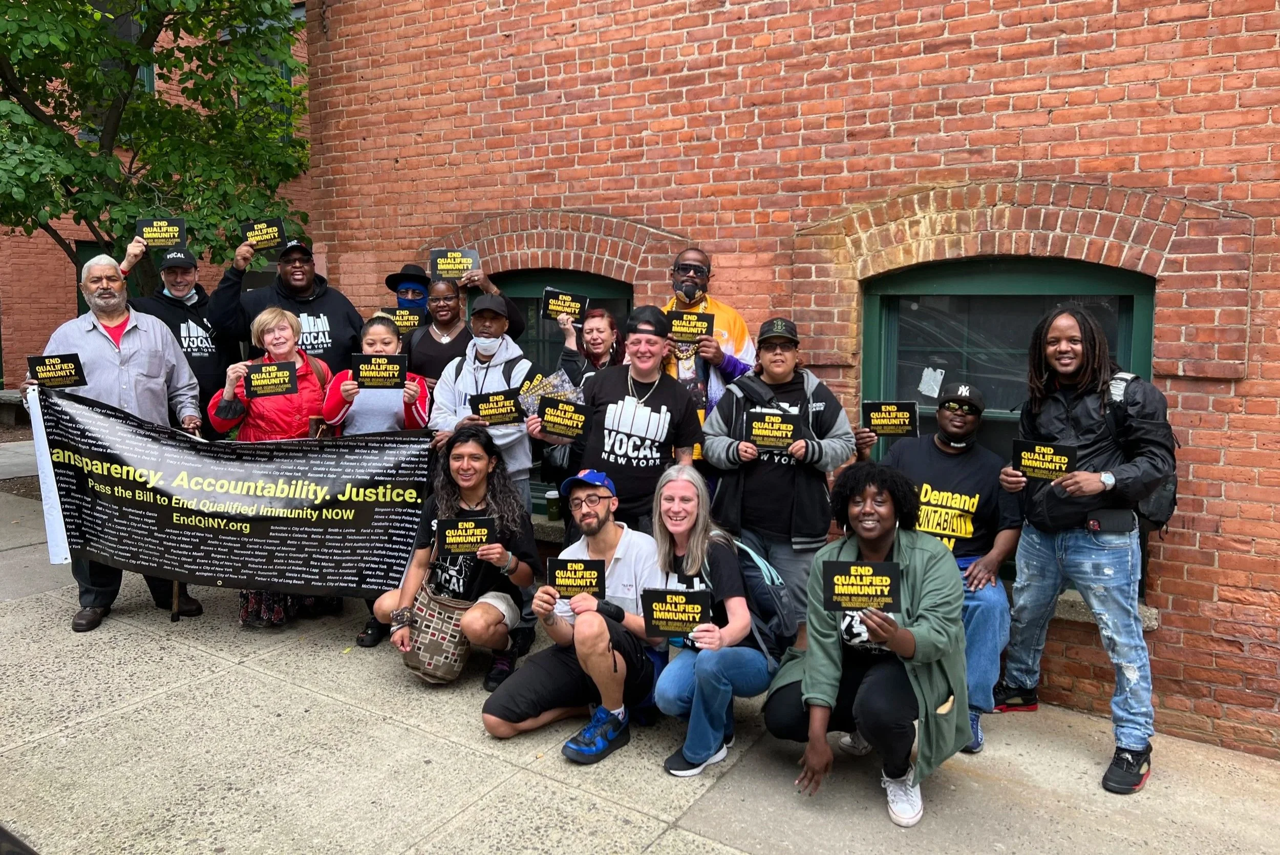 Group of diverse people participating in a rally or protest, holding signs that read "End Qualified Immunity" and a large banner advocating for transparency, accountability, and justice, outdoors on a city sidewalk with brick buildings in the background.
