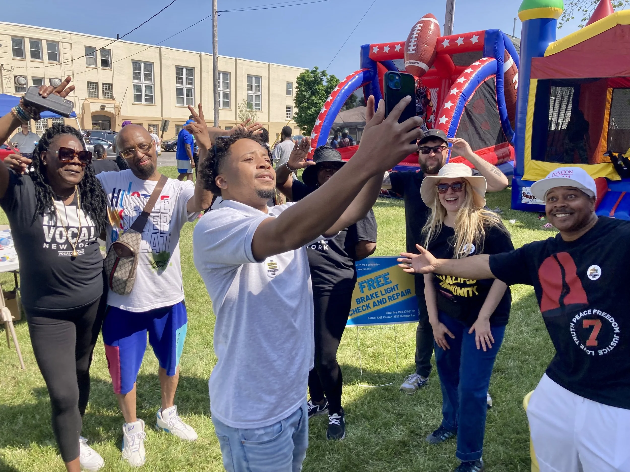A diverse group of people standing on a grassy field at an outdoor event, taking a selfie together with an inflatable bounce house in the background. They are smiling and posing, some making peace signs, under a clear blue sky.