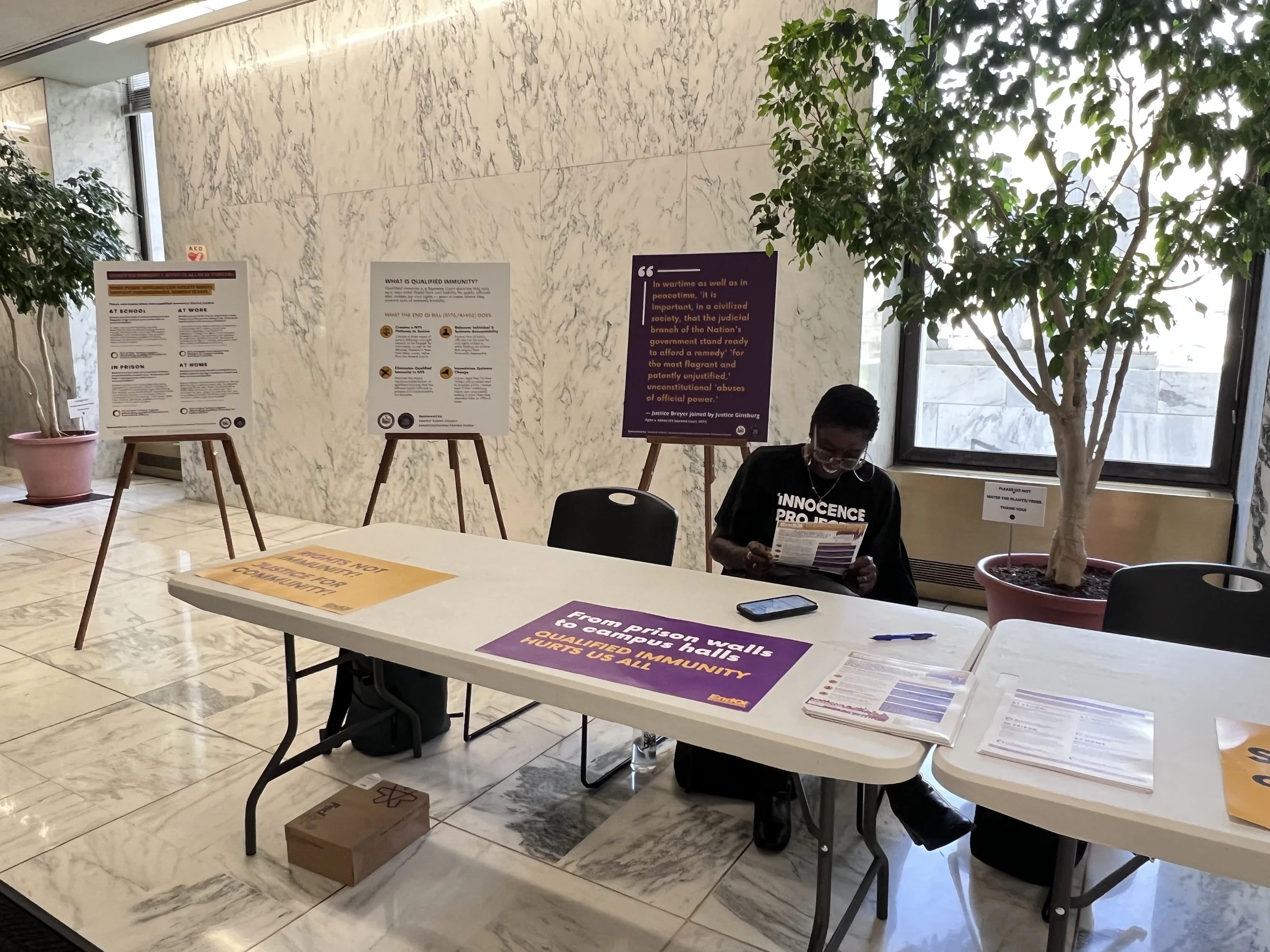 A woman sitting at a table in a lobby or hallway wearing an Innocence Project shirt, reading a pamphlet. There are informational posters on easels behind her about the Bill to End Qualified Immunity in NY, a large potted plant, a window.