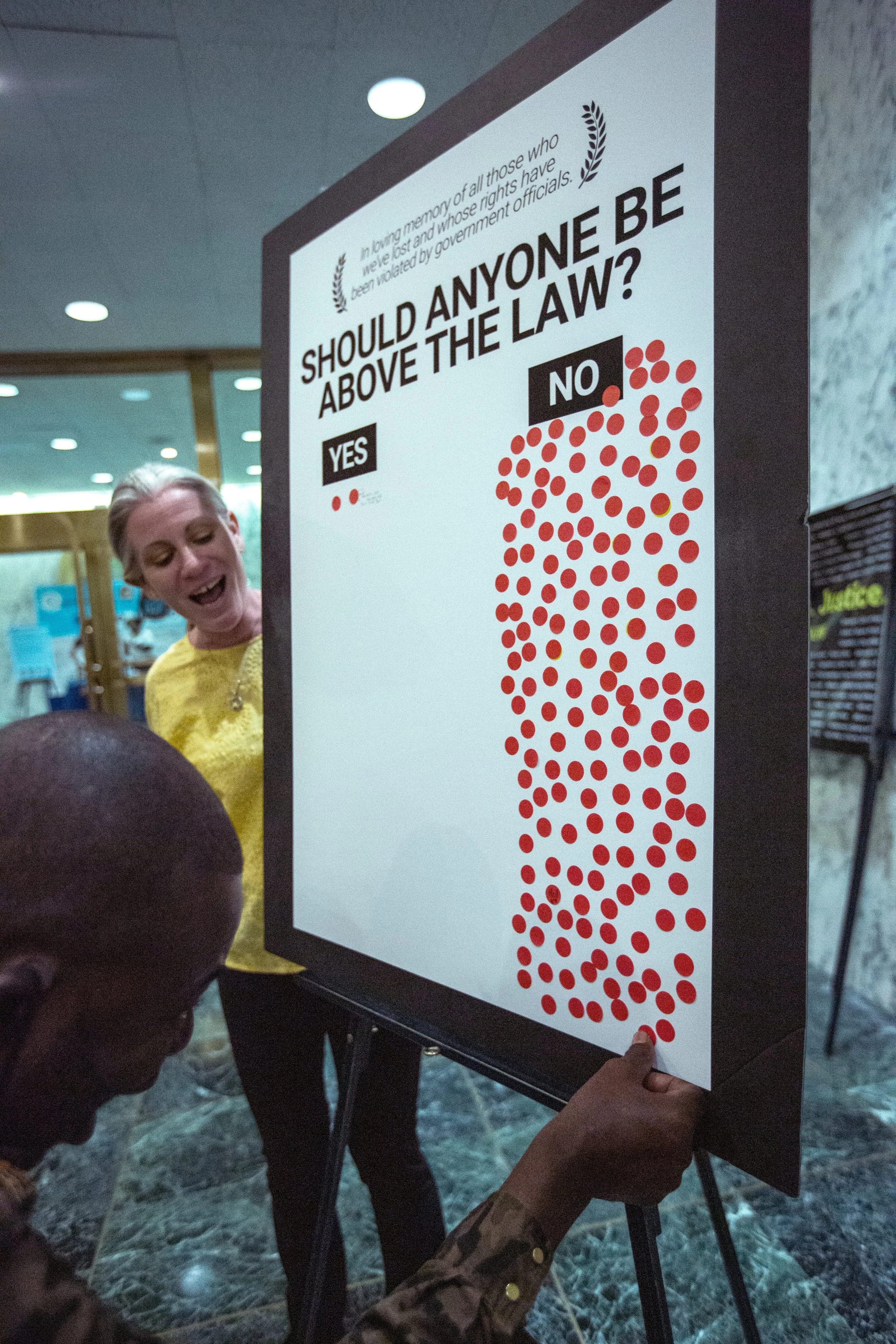 A person holds a poster with "SHOULD ANYONE BE ABOVE THE LAW?" at the top. There are many red stickers under "No" and two under "Yes." Another person is placing a red sticker under "No"