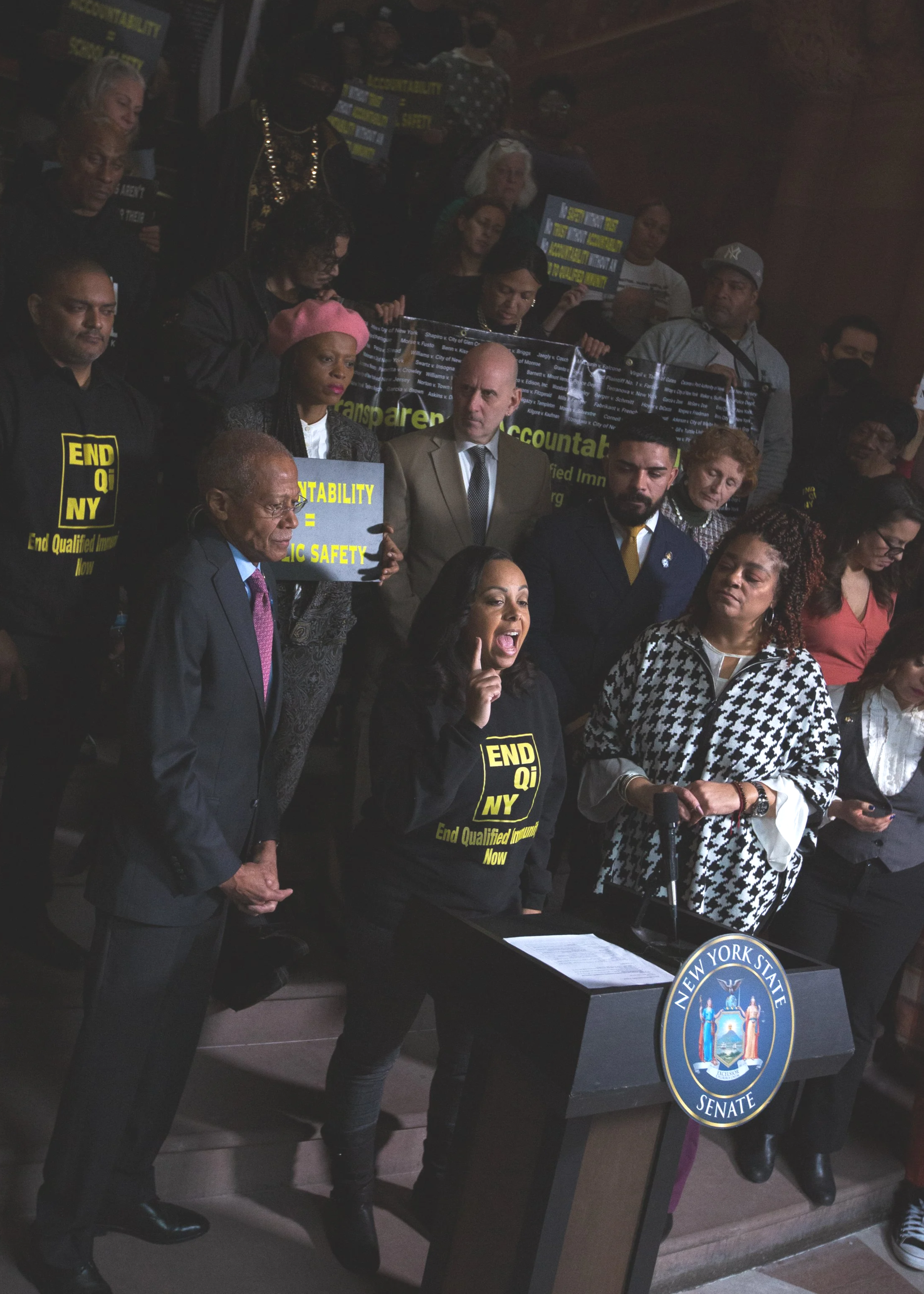 A group of people gathered at a New York State Senate event, some holding protest signs with slogans like 'End Qualified Immunity Now' and 'Accountability = Public Safety.' A woman in front speaks passionately at a podium with the New York State Senate seal.