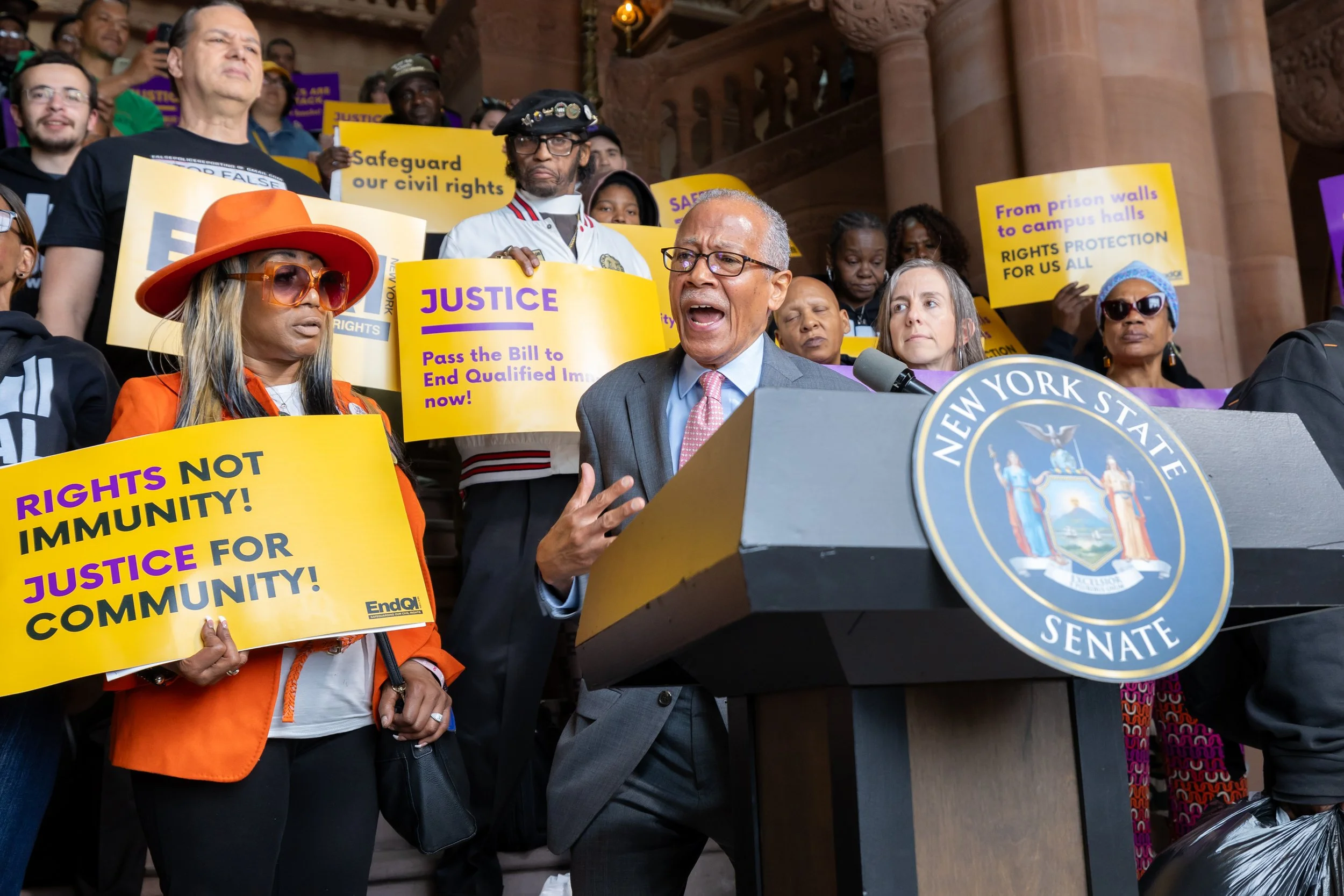 A group of people protesting on the steps of a government building, with some holding yellow signs advocating for civil rights, justice, and community immunity. A man in a suit is speaking at a podium with a seal labeled 'New York State Senate'.