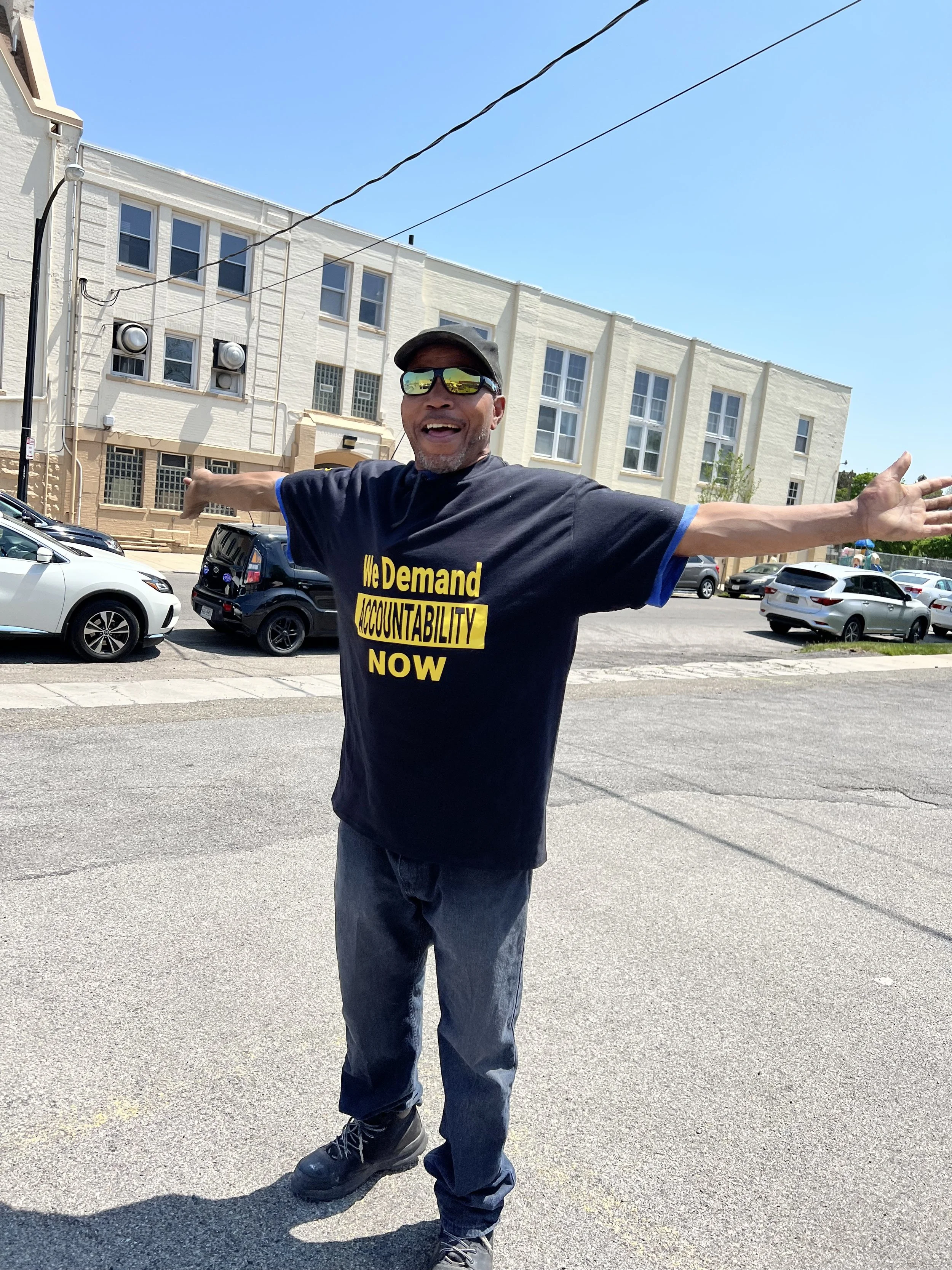 A man wearing a black T-shirt with the message 'We Demand Accountability Now' in yellow and black text, standing outdoors with arms outstretched, smiling, wearing sunglasses, a baseball cap, and jeans, with a background of parked cars and a large white building.