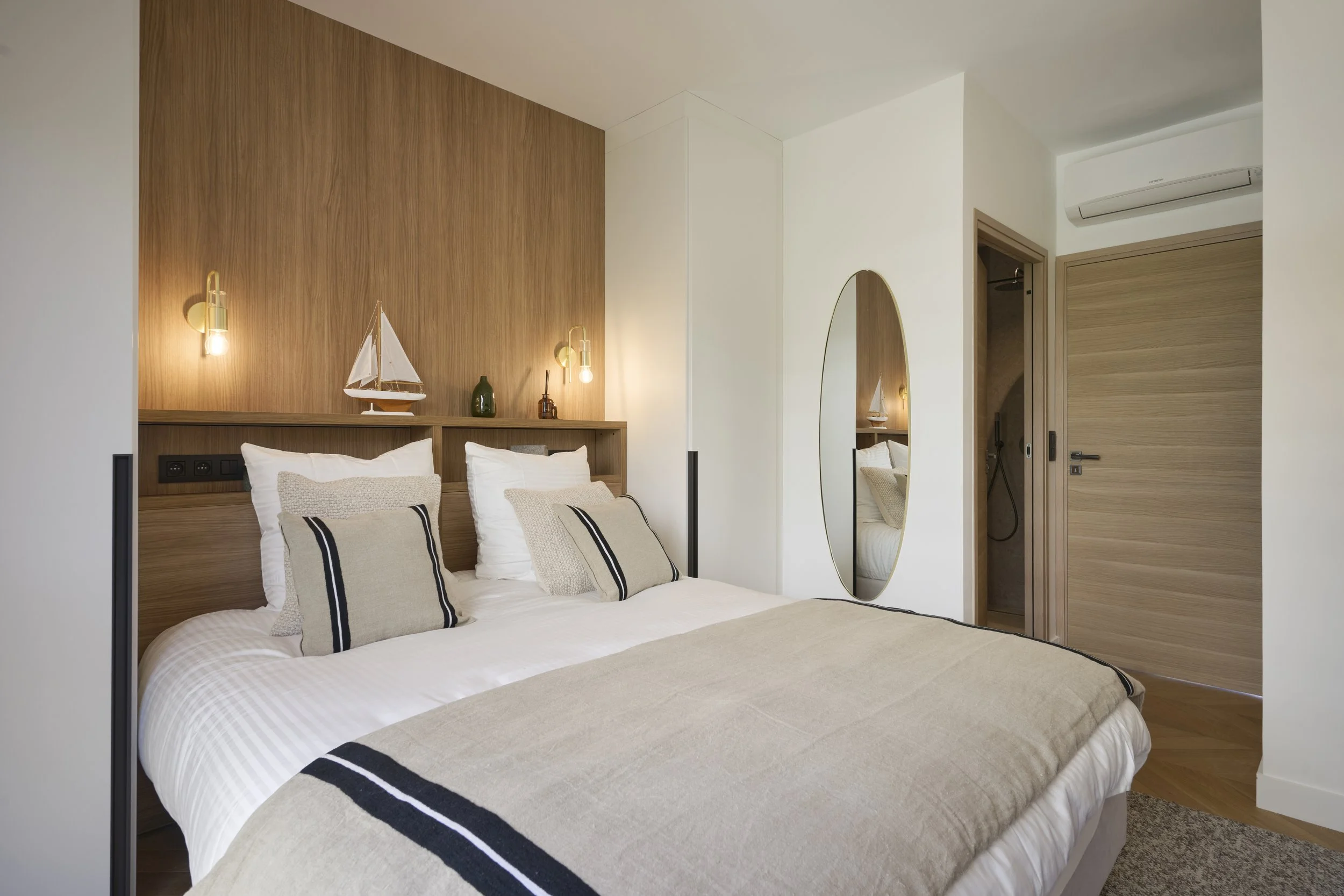 Chambre moderne avec lit double, décoration en bois, coussins beige et blanc, miroir ovale, porte bois, et accents déco nautiques.