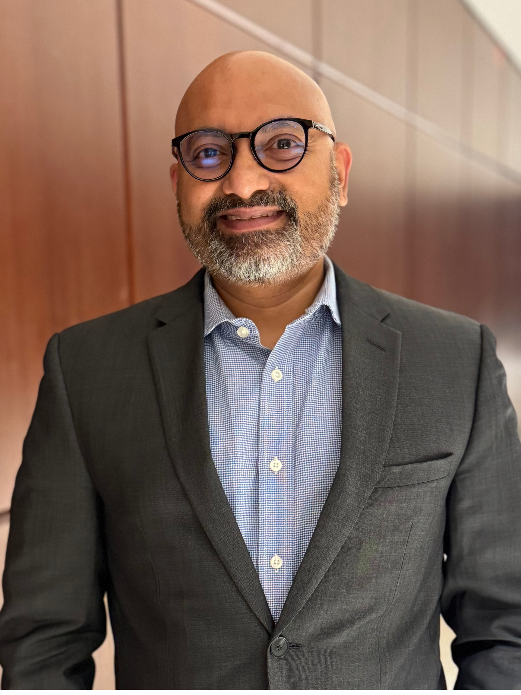 A professional man with glasses, a beard, and a bald head, wearing a dark gray suit and a light blue, checkered dress shirt, smiling in an indoor setting with wooden wall panels in the background.