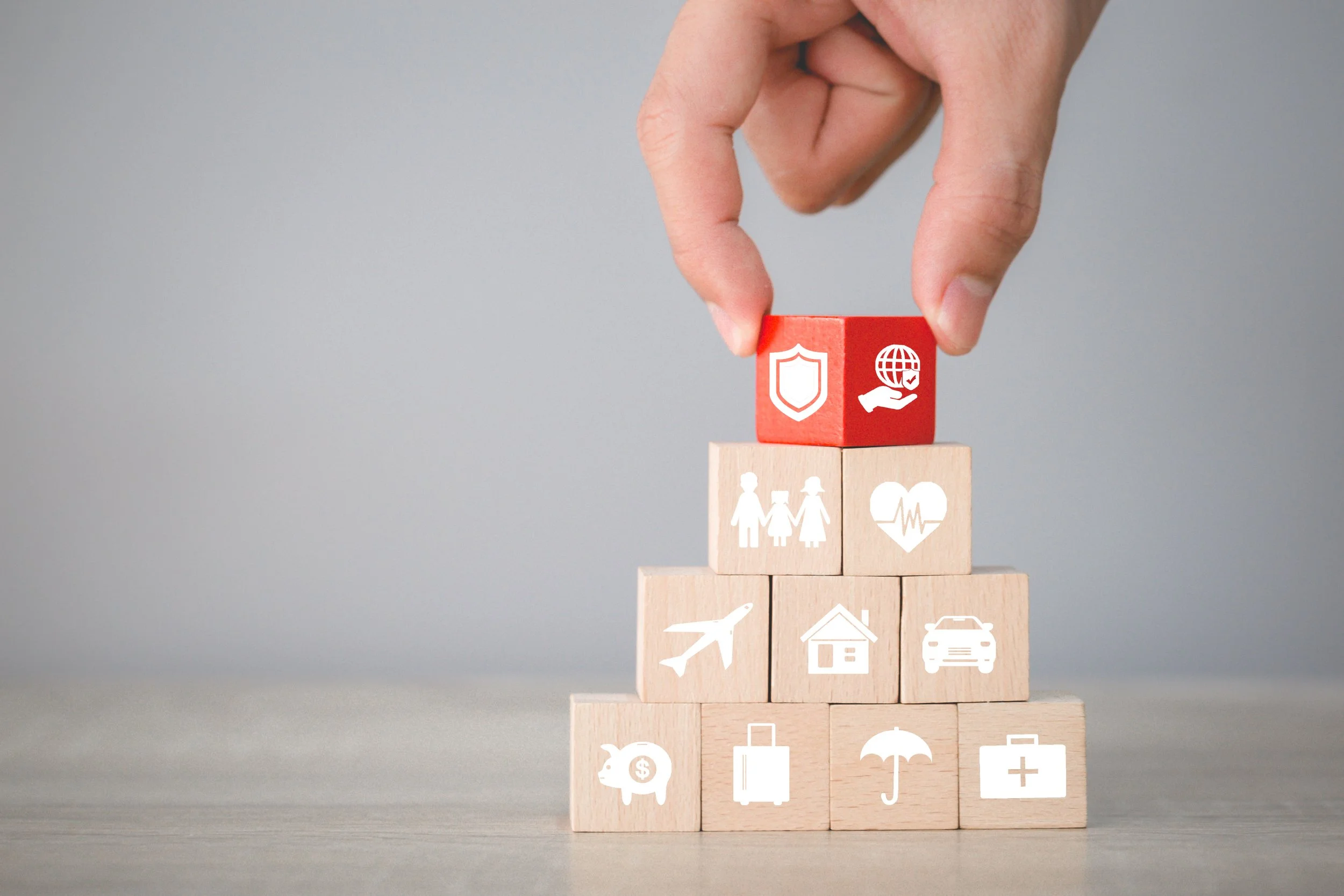Hand legt einen roten Würfel mit einem Schild- und einer Welt-Icon auf einen Turm aus Holzwürfeln mit verschiedenen Icons, darunter Familie, Herz, Flugzeug, Haus, Auto, Schutz, Geldsack, Koffer, Regenschirm und Medizinbox.