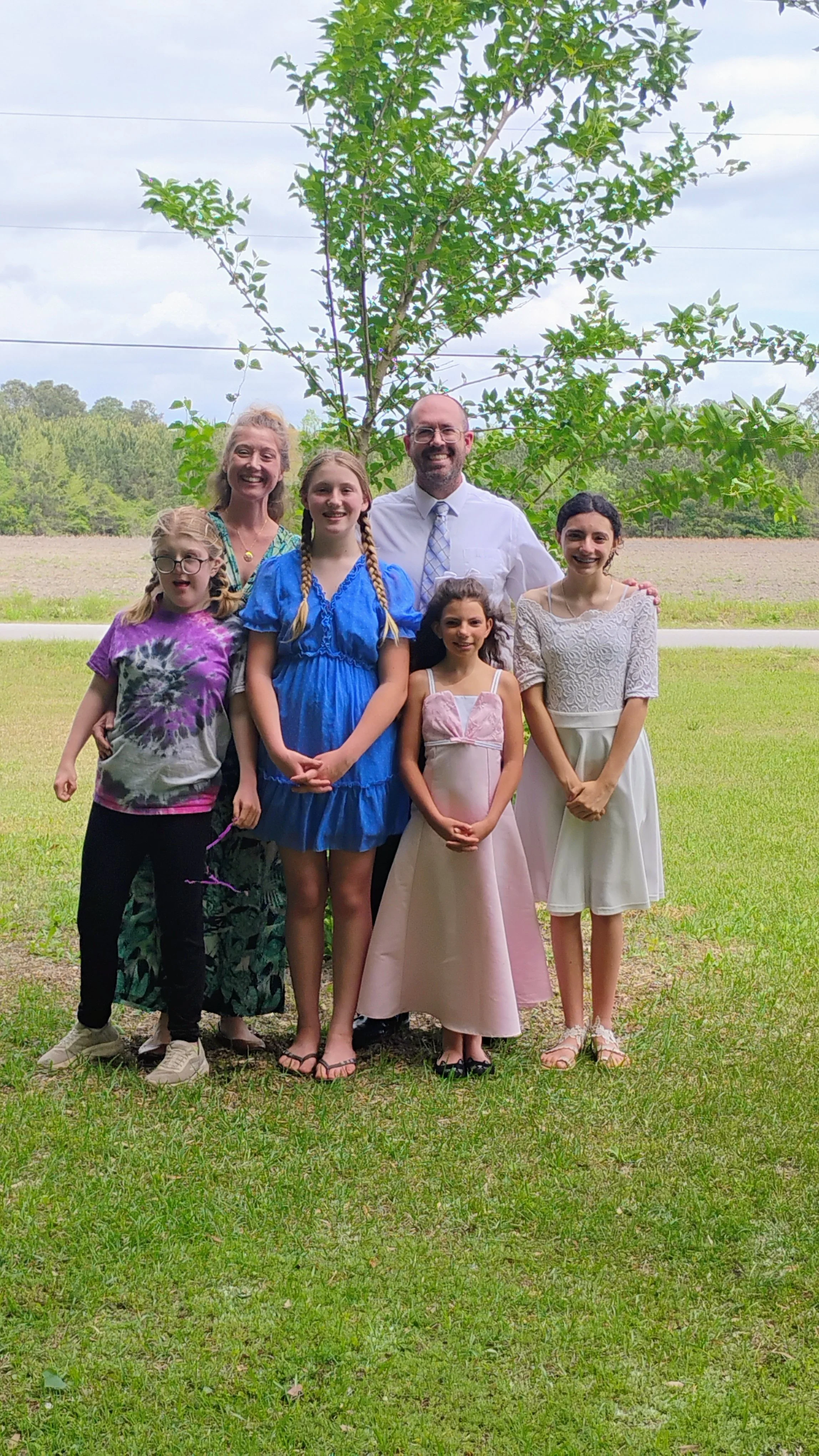 Group photo of six people, including children and adults, standing on grass in front of a tree outdoors. They are smiling, with some dressed in casual clothing and others in dresses.