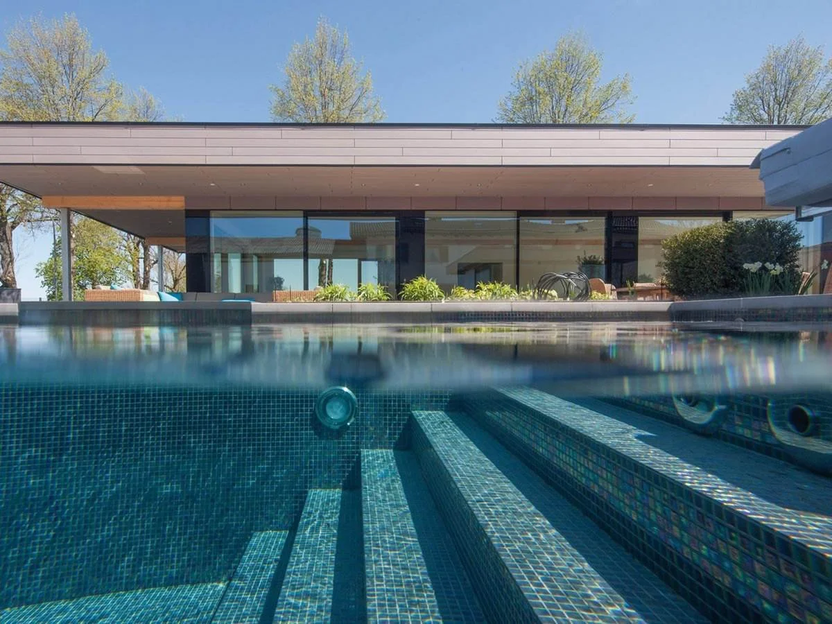 Piscina con mosaico azzurro e casa moderna sullo sfondo, con alberi e cielo azzurro.