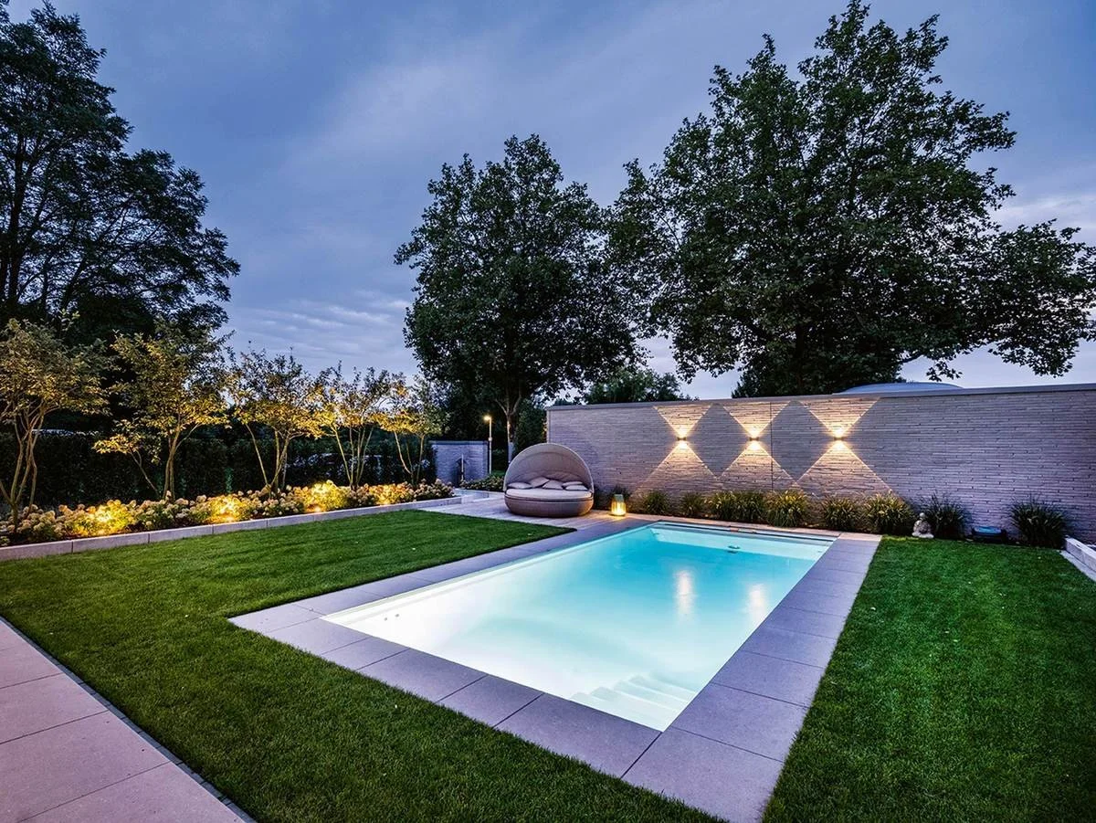 Piscina illuminata in un giardino con prato verde, luci decorative sulla parete, e alberi al tramonto.