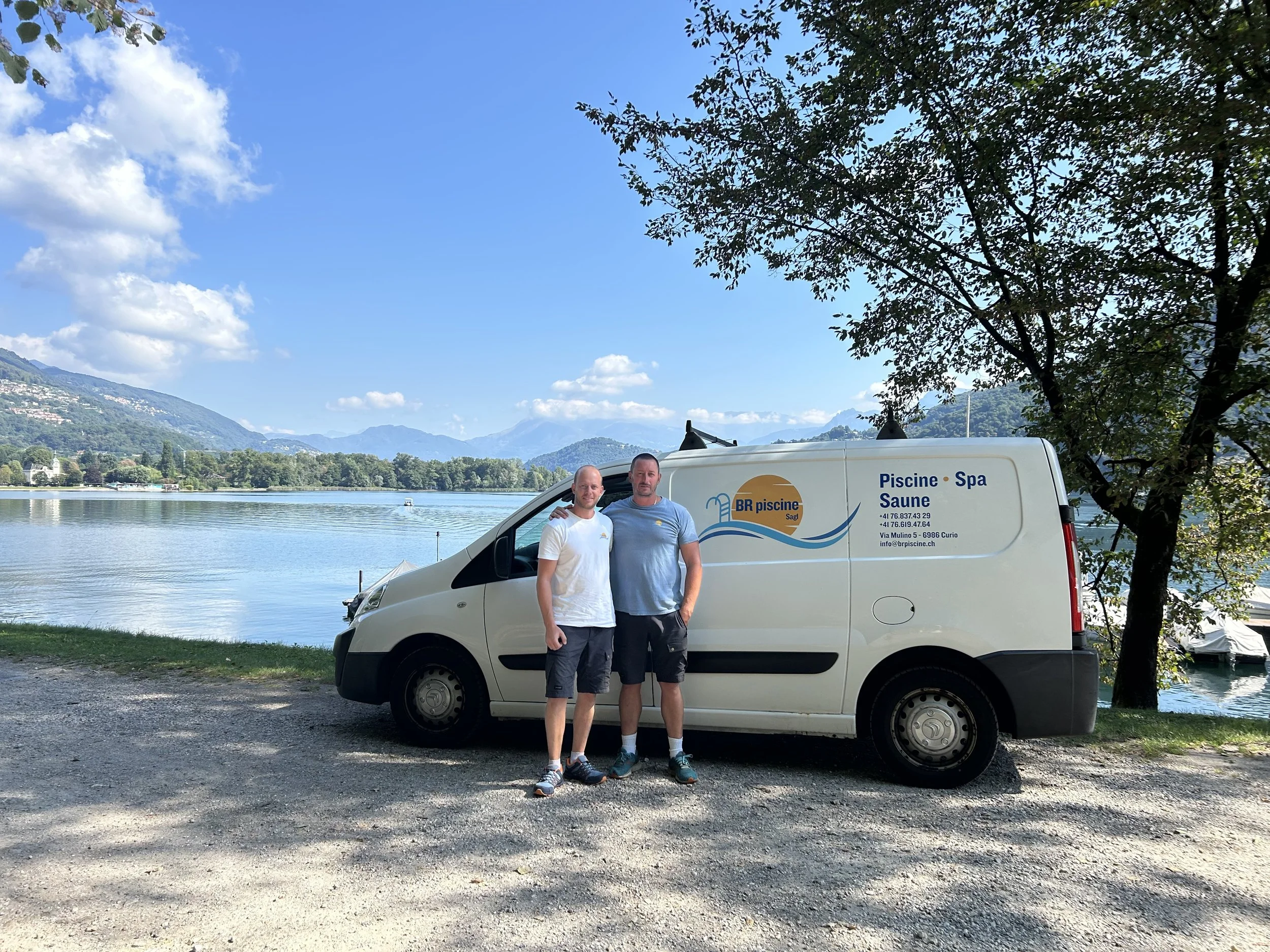 Due uomini davanti a un'auto bianca con un lago e montagne sullo sfondo, piantagioni di alberi e un cielo soleggiato.