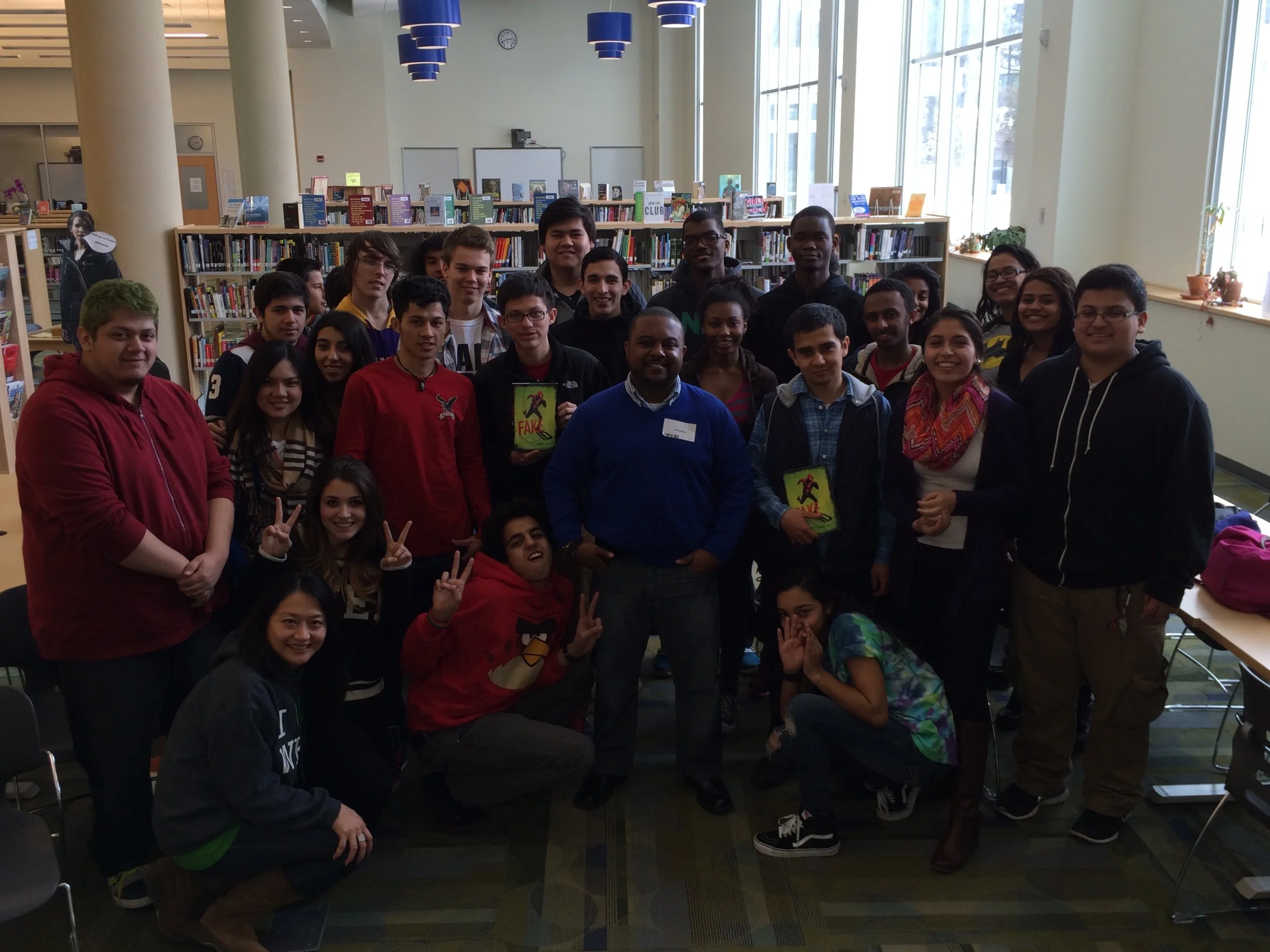 Group of young people and author Lamar Giles posing for a photo inside a library, some holding books, with bookshelves and large windows in the background.
