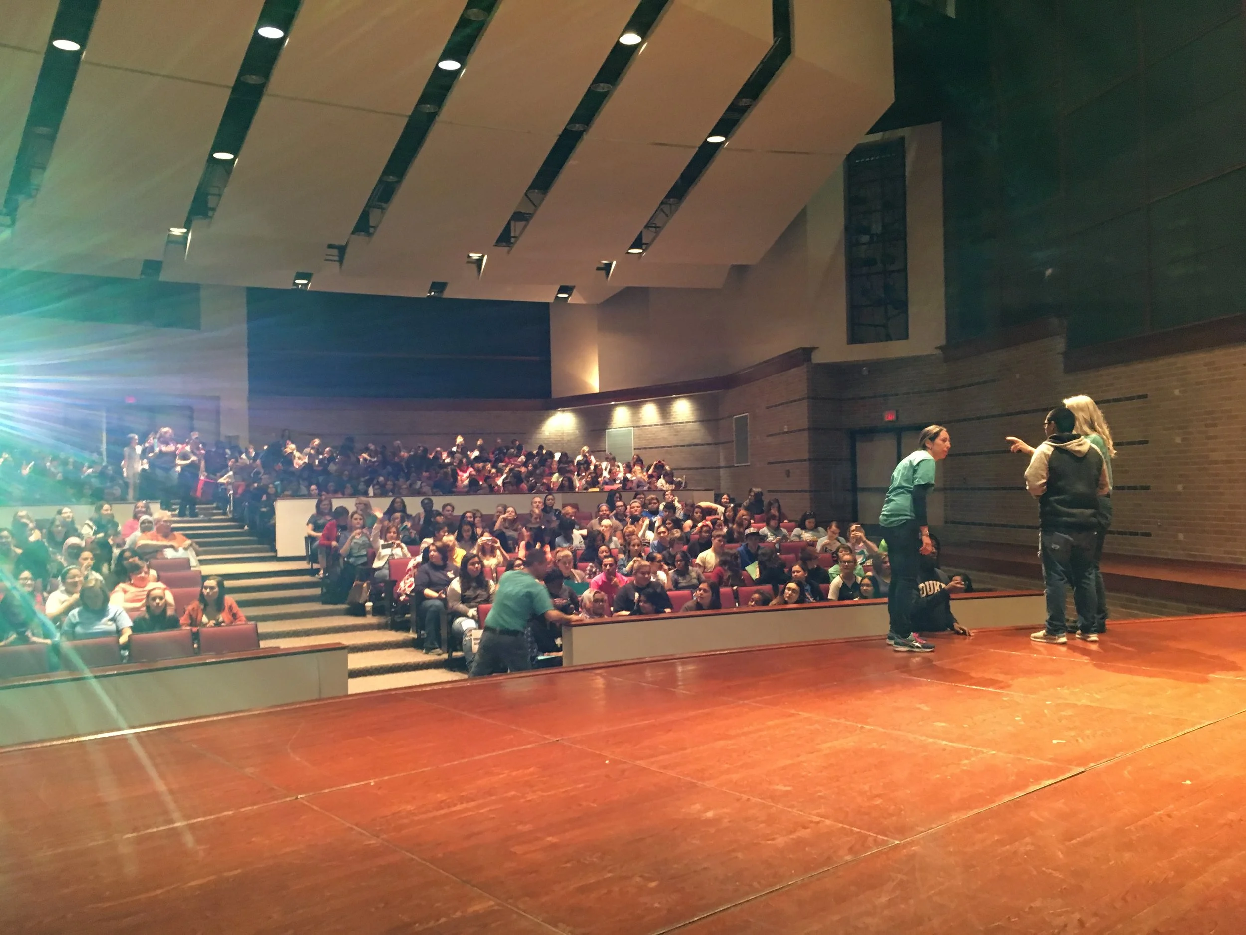 A large auditorium filled with students seated in rows of chairs, watching a stage where three women are engaged in conversation.