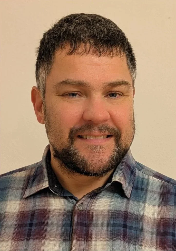 Ein Mann mit dunklem, kurzem Haar und Bart, trägt ein kariertes Hemd und lächelt vor neutralem Hintergrund.