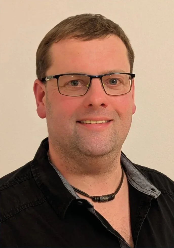 Porträt eines Mannes mit Brille, dunklem Hemd und einem schwarzen Halsband vor neutralem Hintergrund.