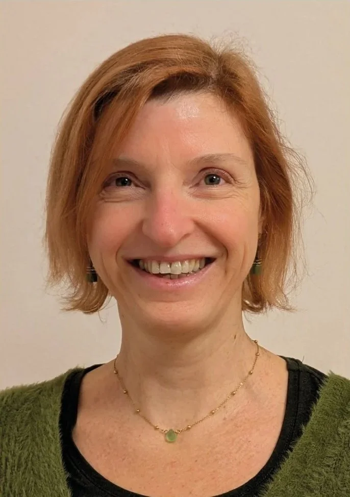 Lächelnde Frau mit kurzen roten Haaren, trägt eine grüne Jacke, schwarze Oberteil und eine goldene Halskette mit grünem Anhänger.