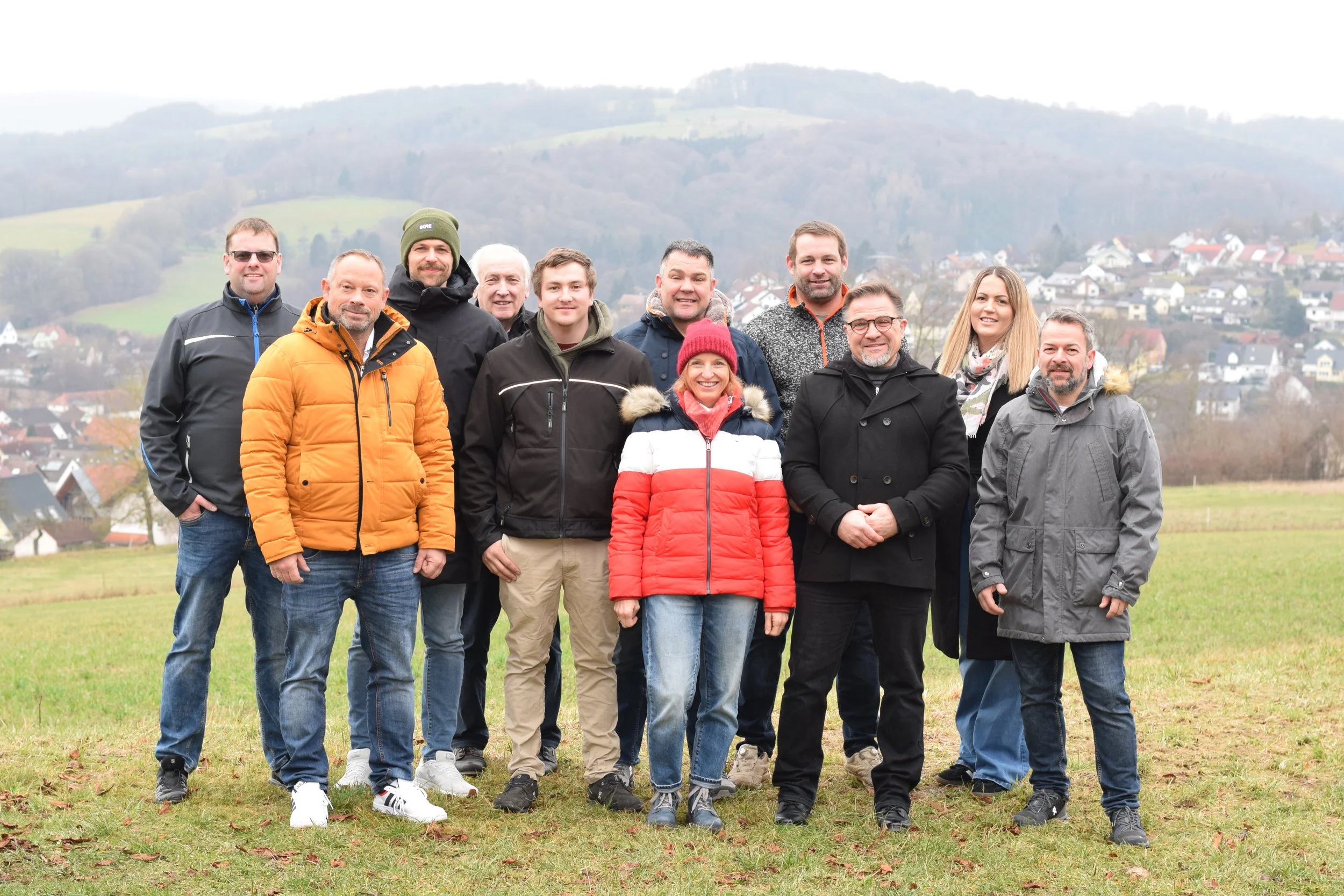 Gruppe von 13 Personen im Freien auf einer Wiese mit Hügeln und Häuser im Hintergrund, alle tragen Jacken für das kalte Wetter.