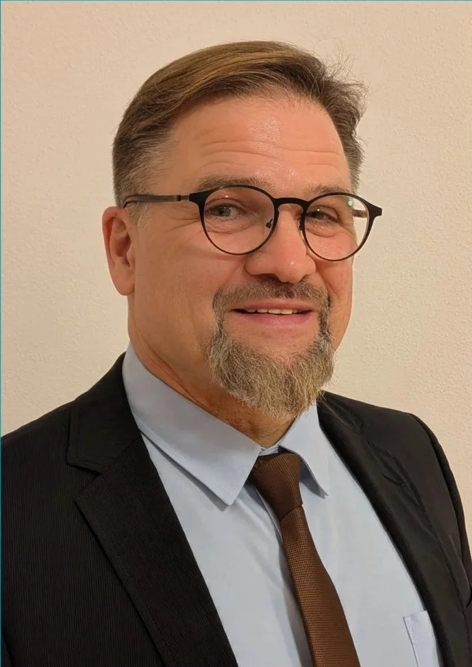 Ein Mann trägt einen schwarzen Anzug, ein hellblaues Hemd, eine braune Krawatte und runde Brillen, steht vor einer hellen Wand und lächelt leicht.