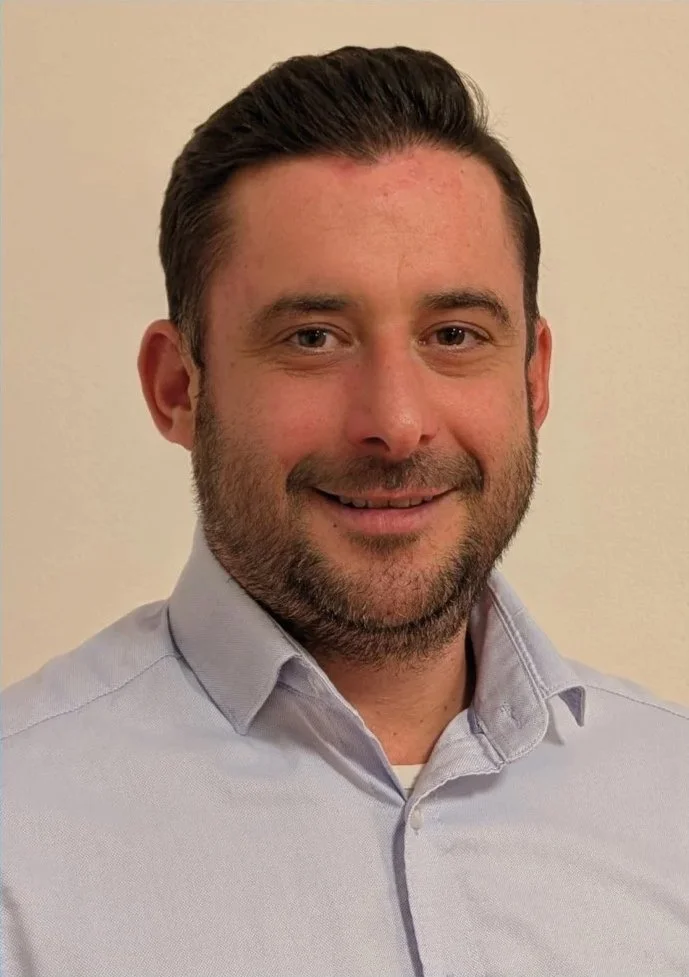 Ein Mann mit dunklen Haaren, Bart und einem hellblauen Hemd, lächelnd vor einer hellen Wand.