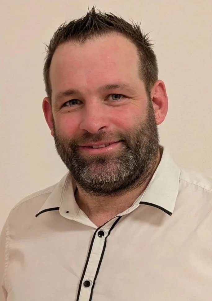 Ein lachender Mann mit Bart in einem weißen Hemd gegen eine helle Wand.